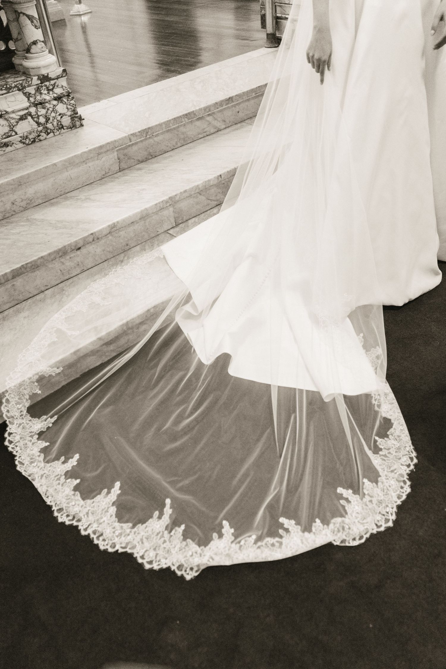 Black and white close-up of bride's cathedral-length lace-trimmed wedding veil trailing on church floor.