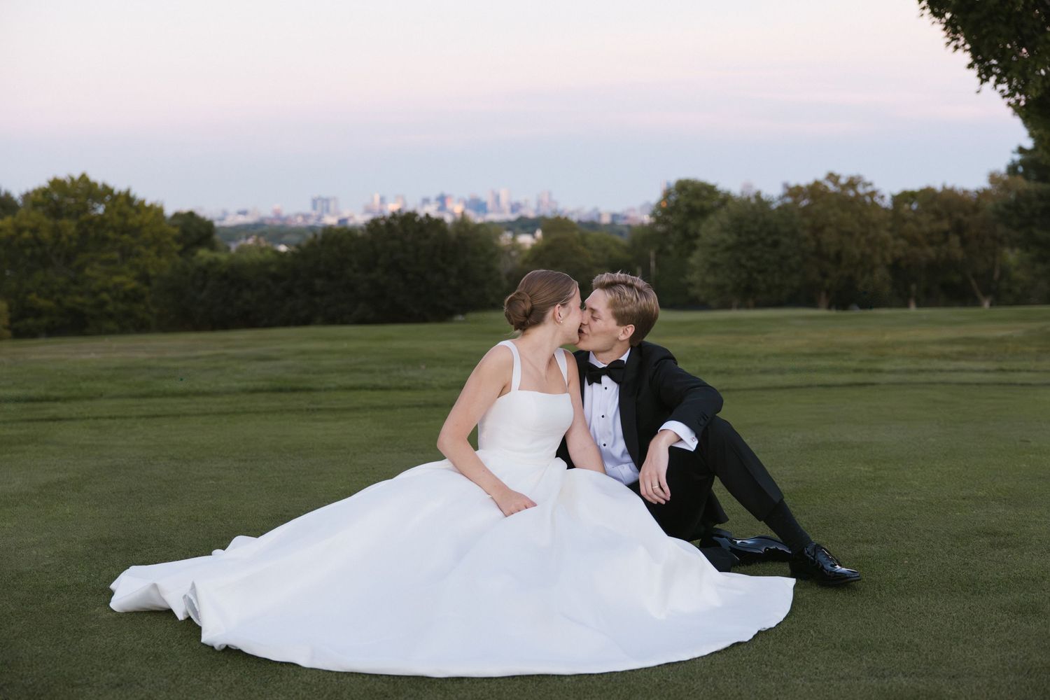 Bride and groom share a kiss while sitting on a golf course lawn at dusk.