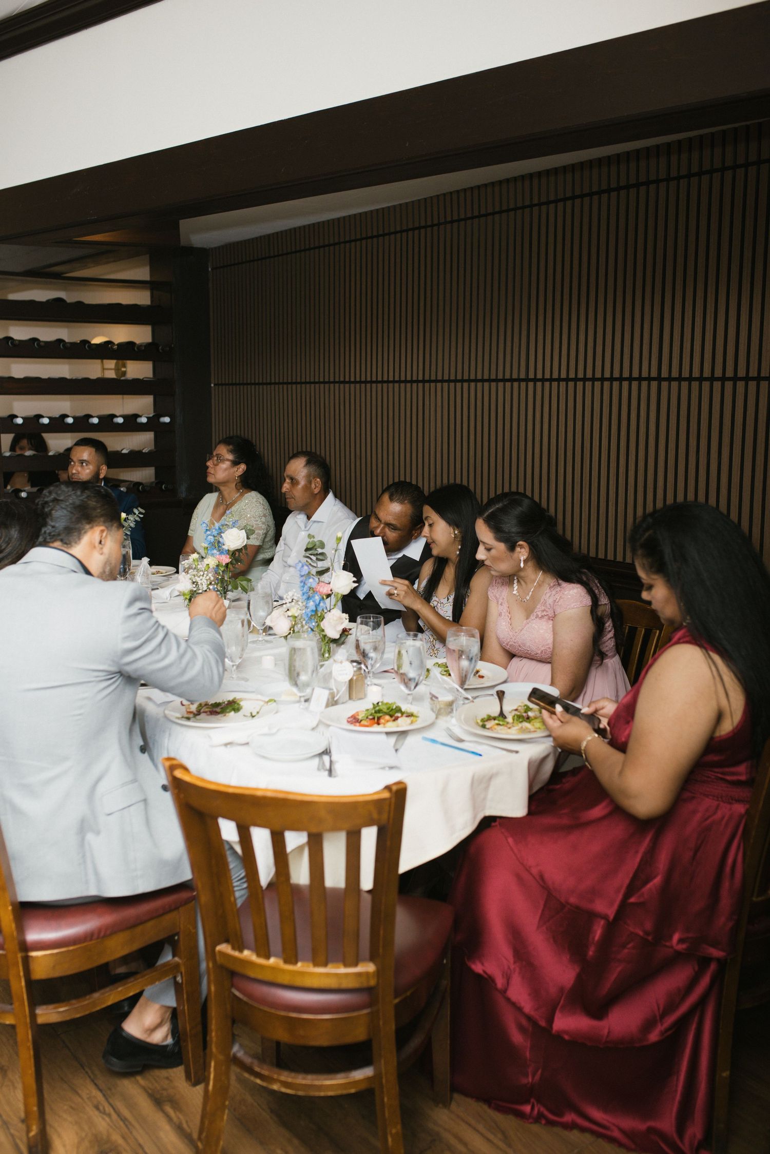 Wedding reception guests seated at round dining table enjoying dinner and speeches at elegant venue.