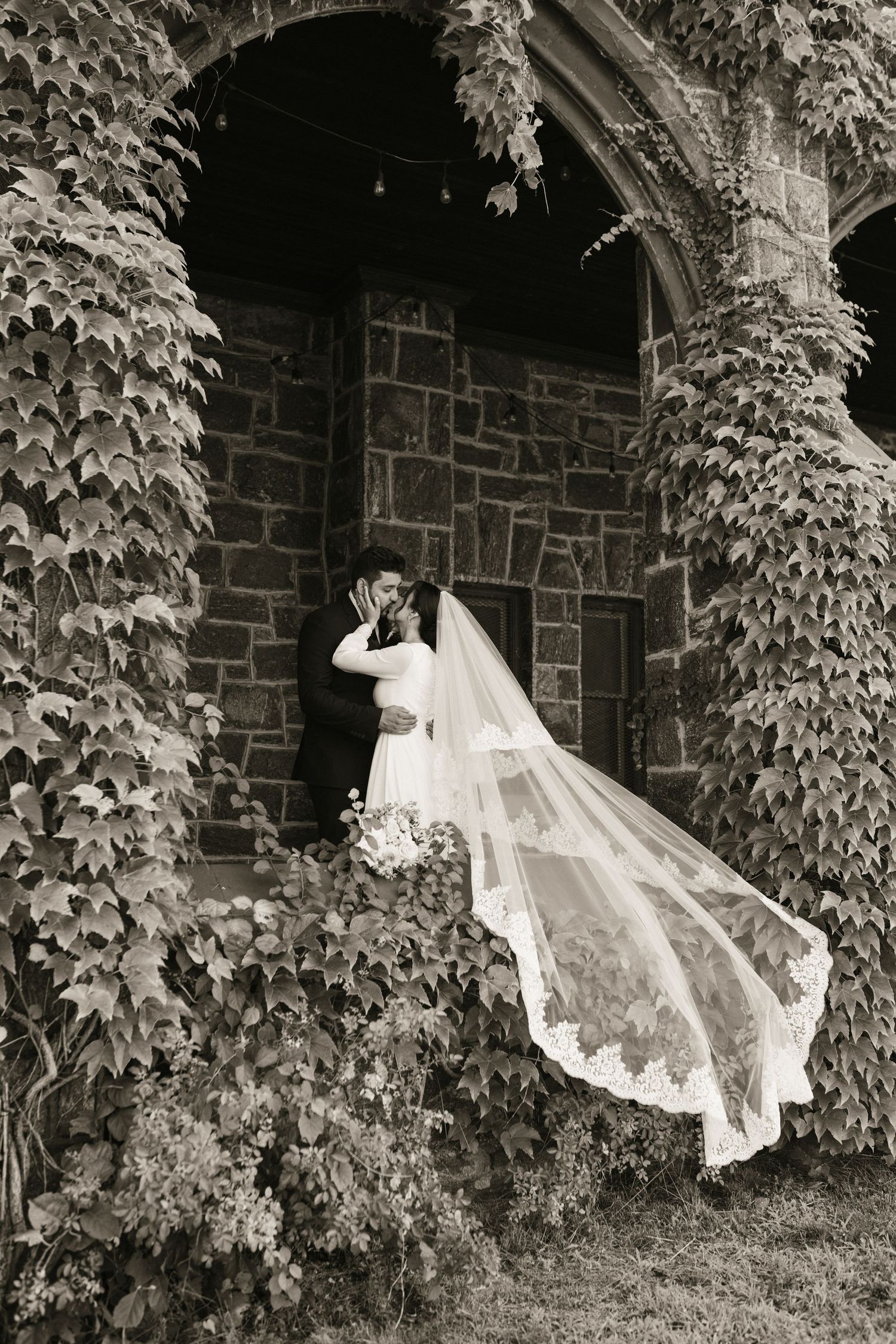Bride and groom share a kiss framed by ivy-covered stone archway in black and white photo.