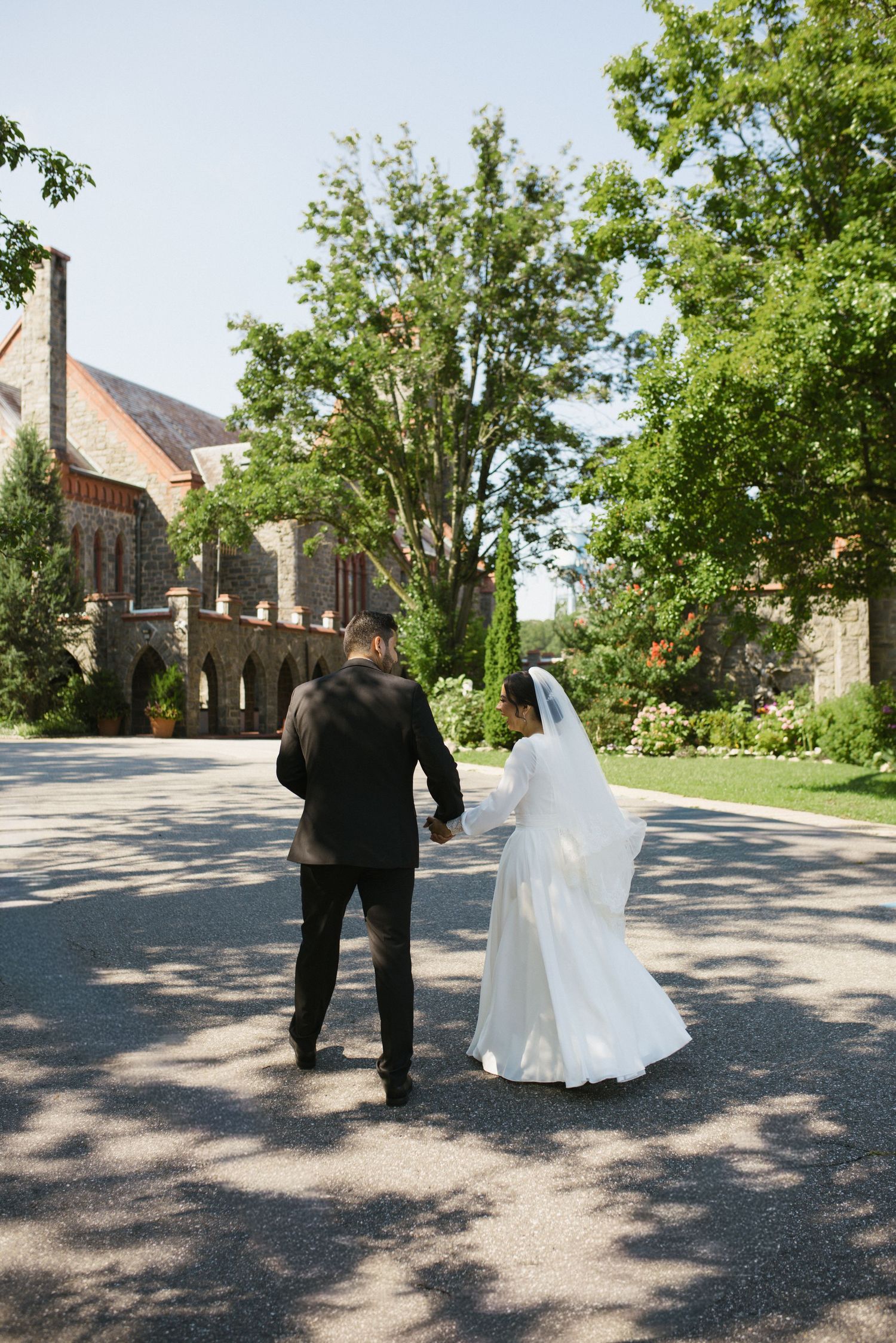 Bride and groom walk hand-in-hand along a tree-lined path at a historic stone estate.