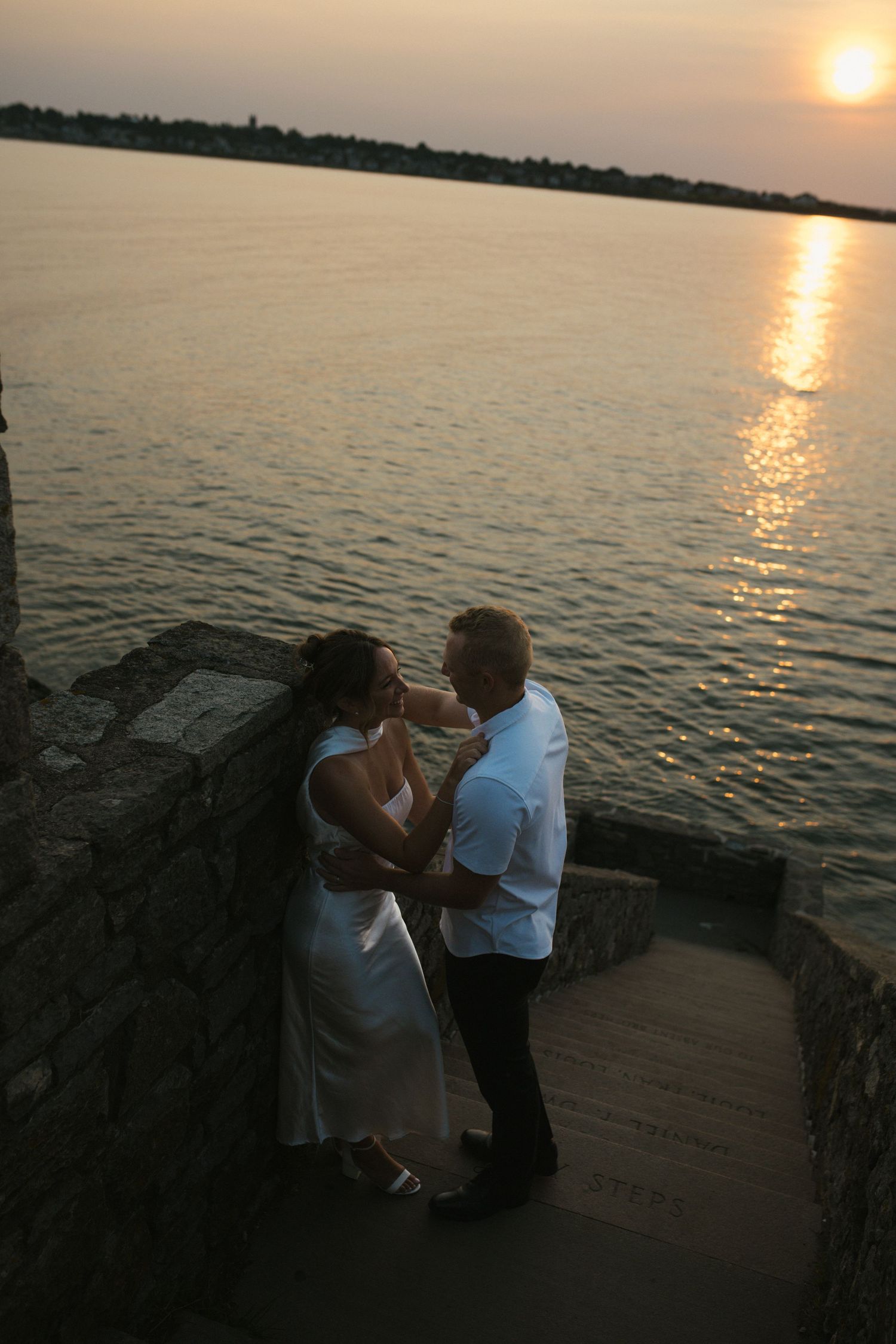 Couple in white embracing at sunset by the water on stone steps, romantic golden hour scene.