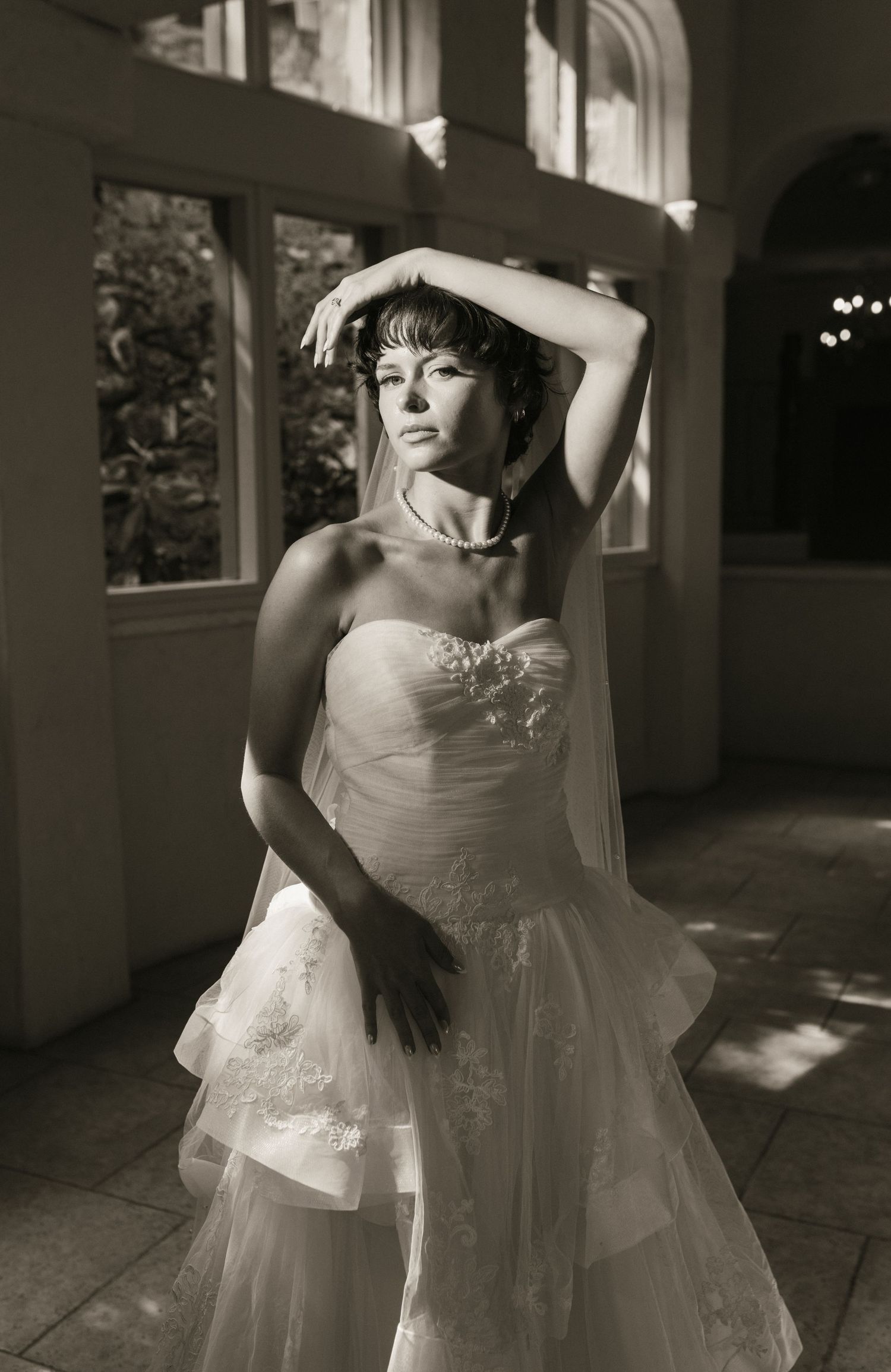 Black and white photo of bride in strapless lace wedding gown posing dramatically in sunlit venue hallway.