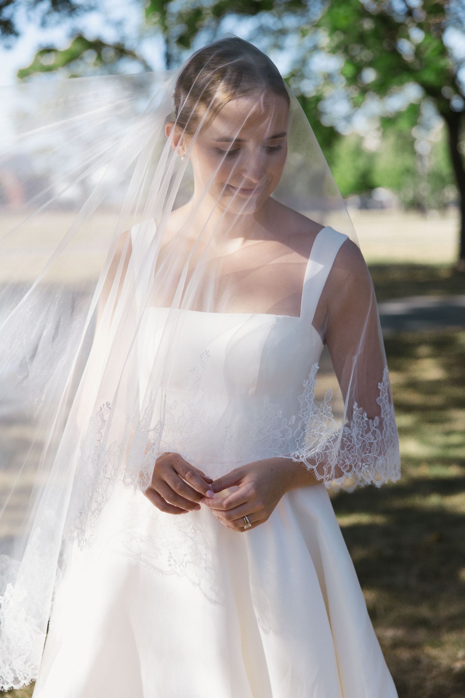 Elegant bride in white gown and lace-trimmed veil standing outdoors under shaded trees.