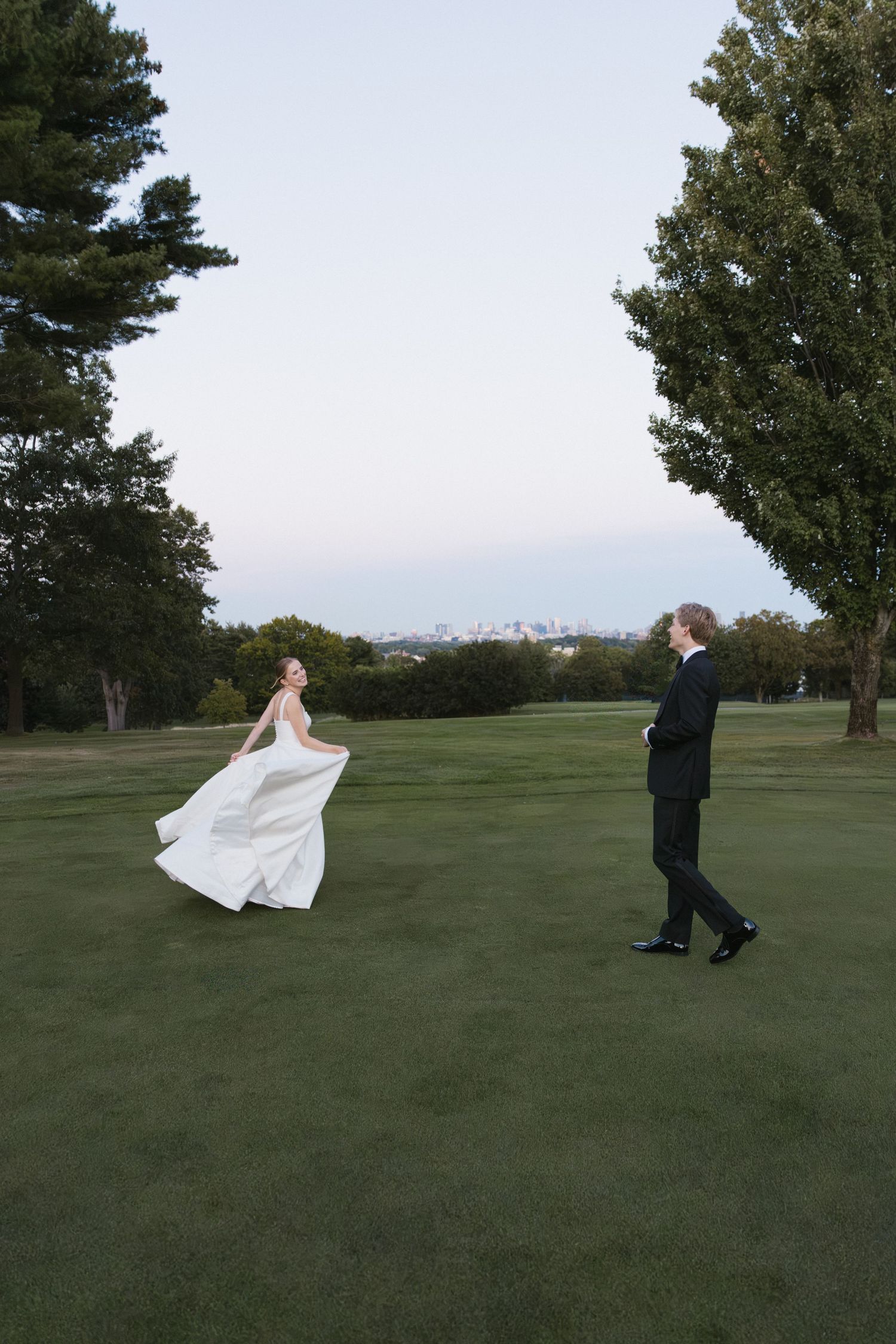 Bride in flowing white gown twirls for groom on lush green golf course at dusk.