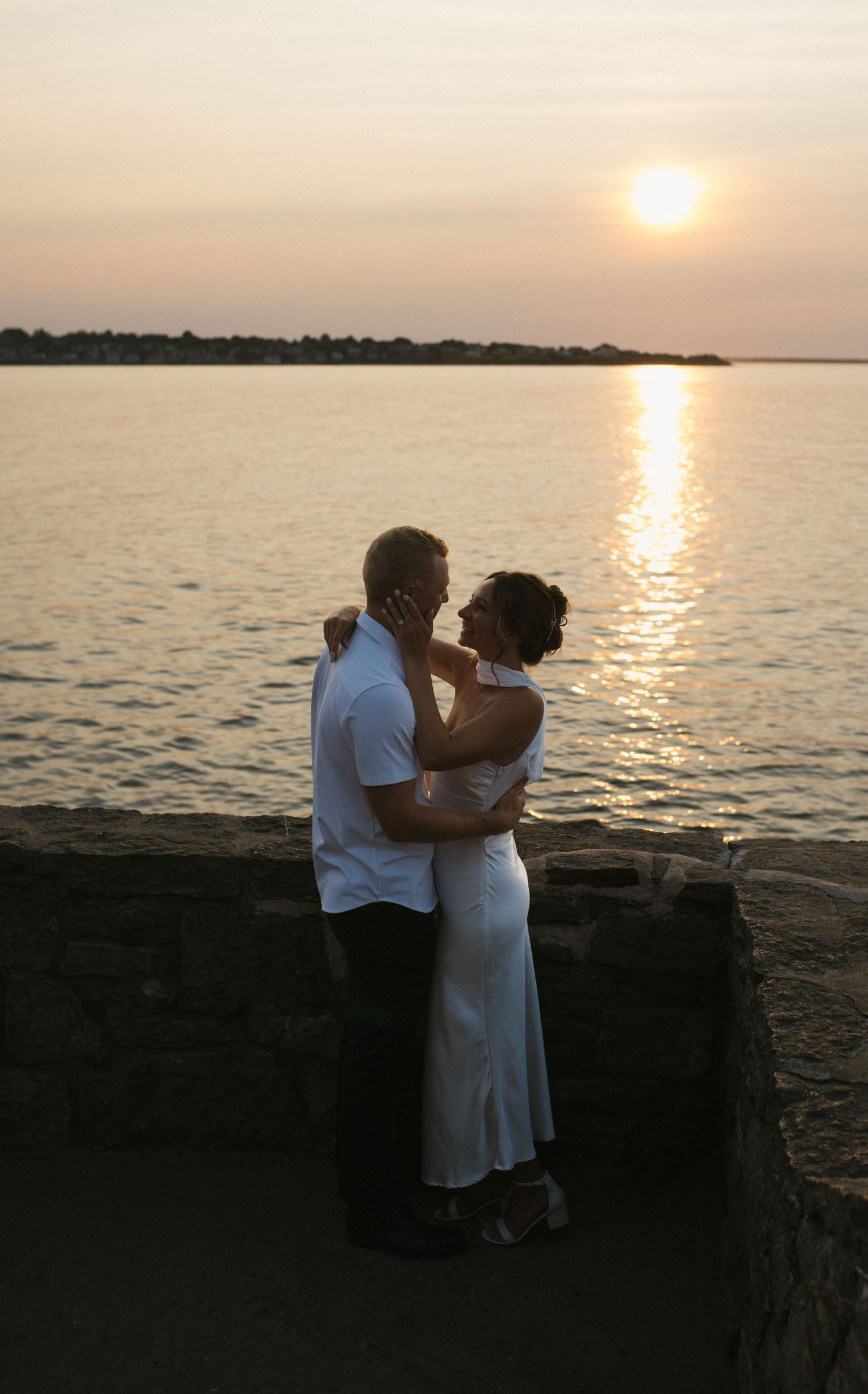 Romantic couple embracing at sunset by the water, woman in white dress, man in white shirt.