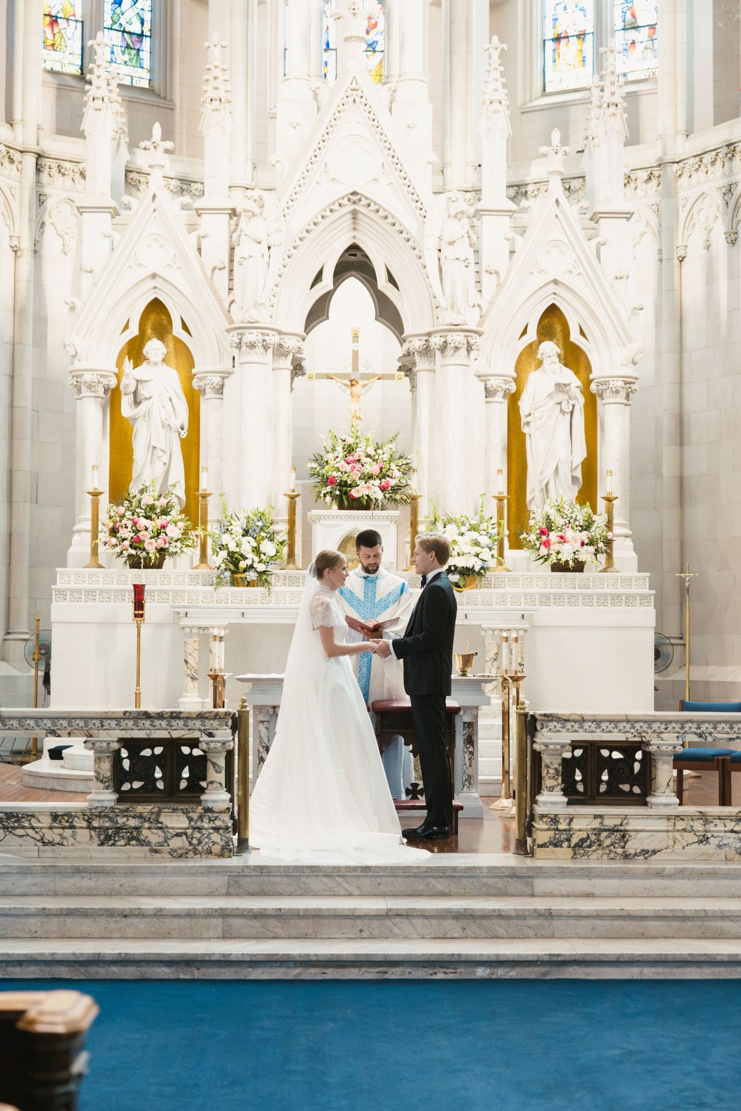 Bride and groom exchange vows at ornate white Gothic church altar with floral arrangements and stained glass windows.