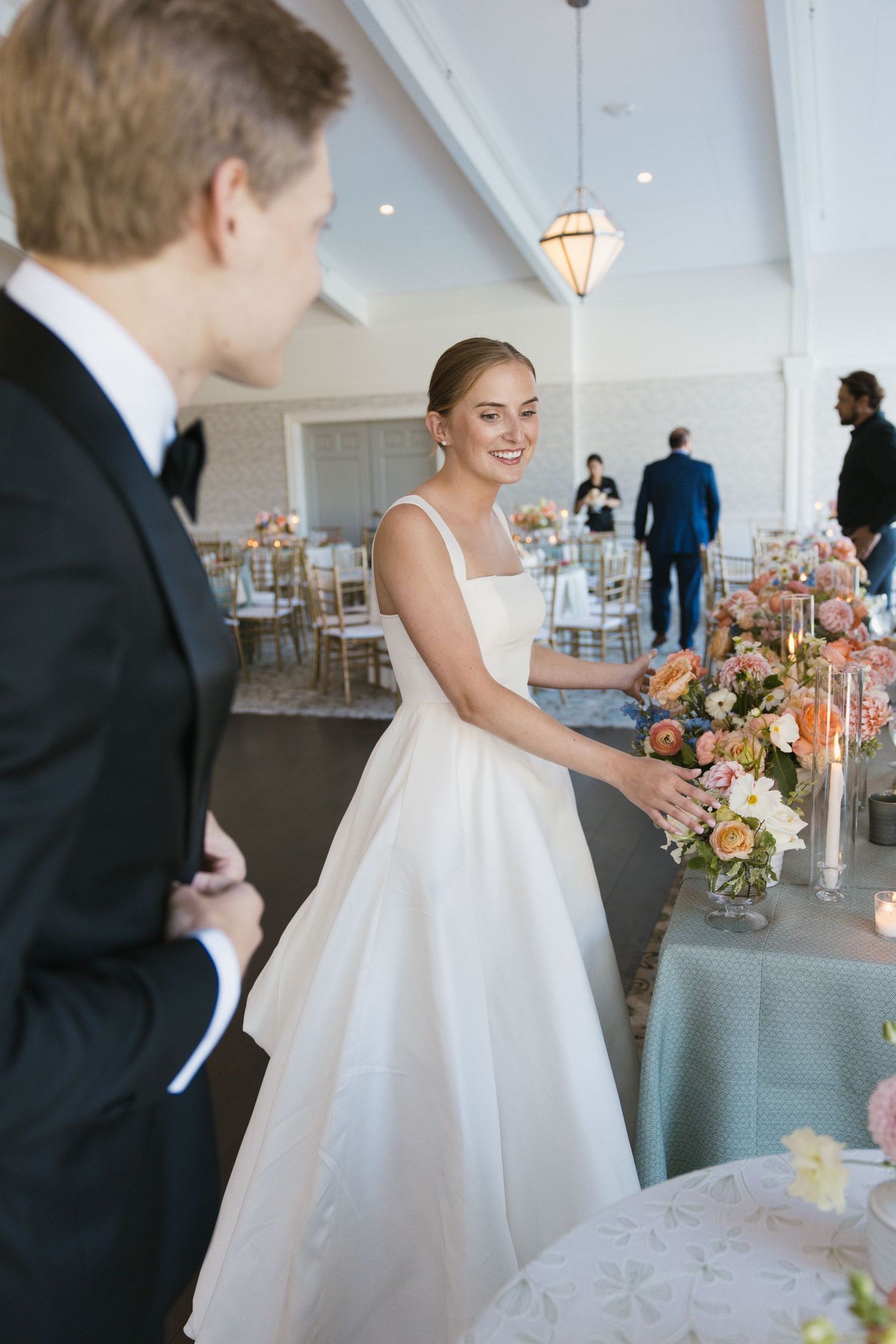 Smiling bride in white gown arranges floral centerpieces at elegantly decorated wedding reception tables.