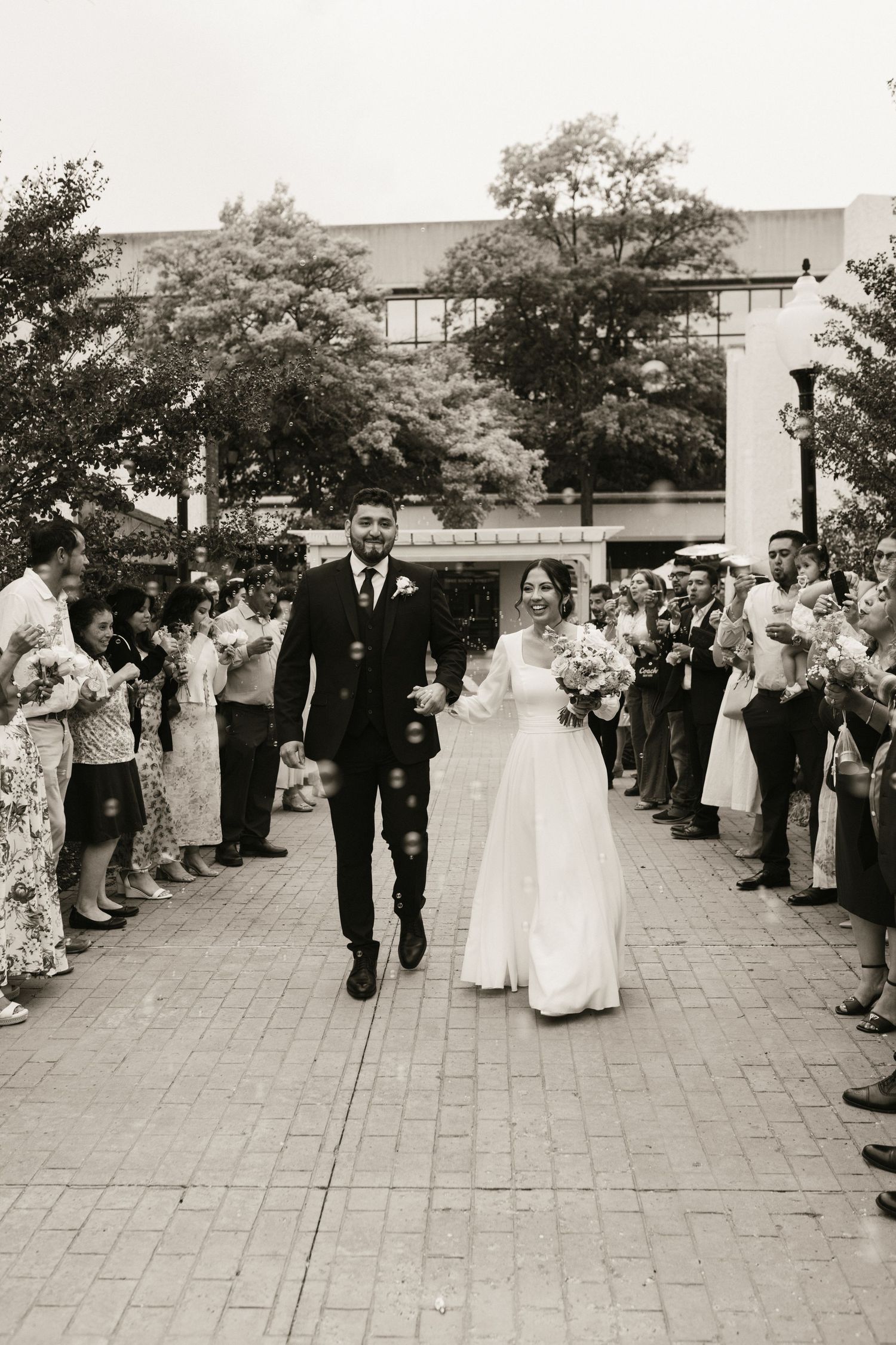 Bride and groom exit church in black and white photo as guests line path blowing bubbles.