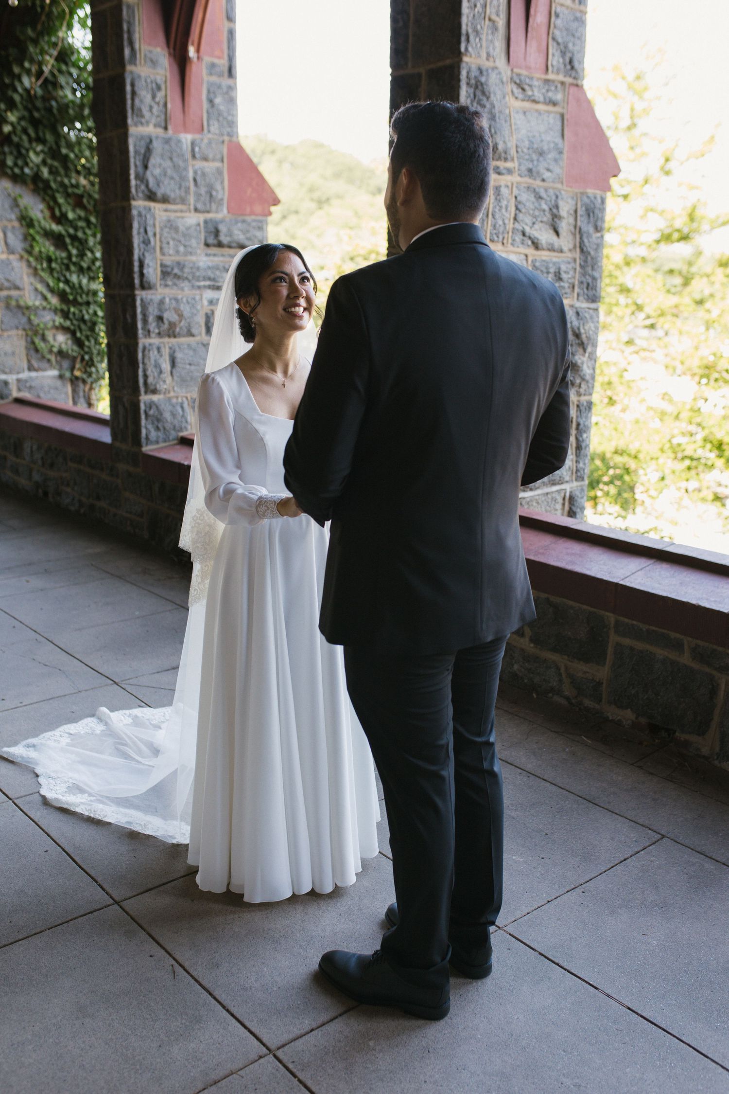 Smiling bride in white gown holds grooms hands during first look at stone castle venue.