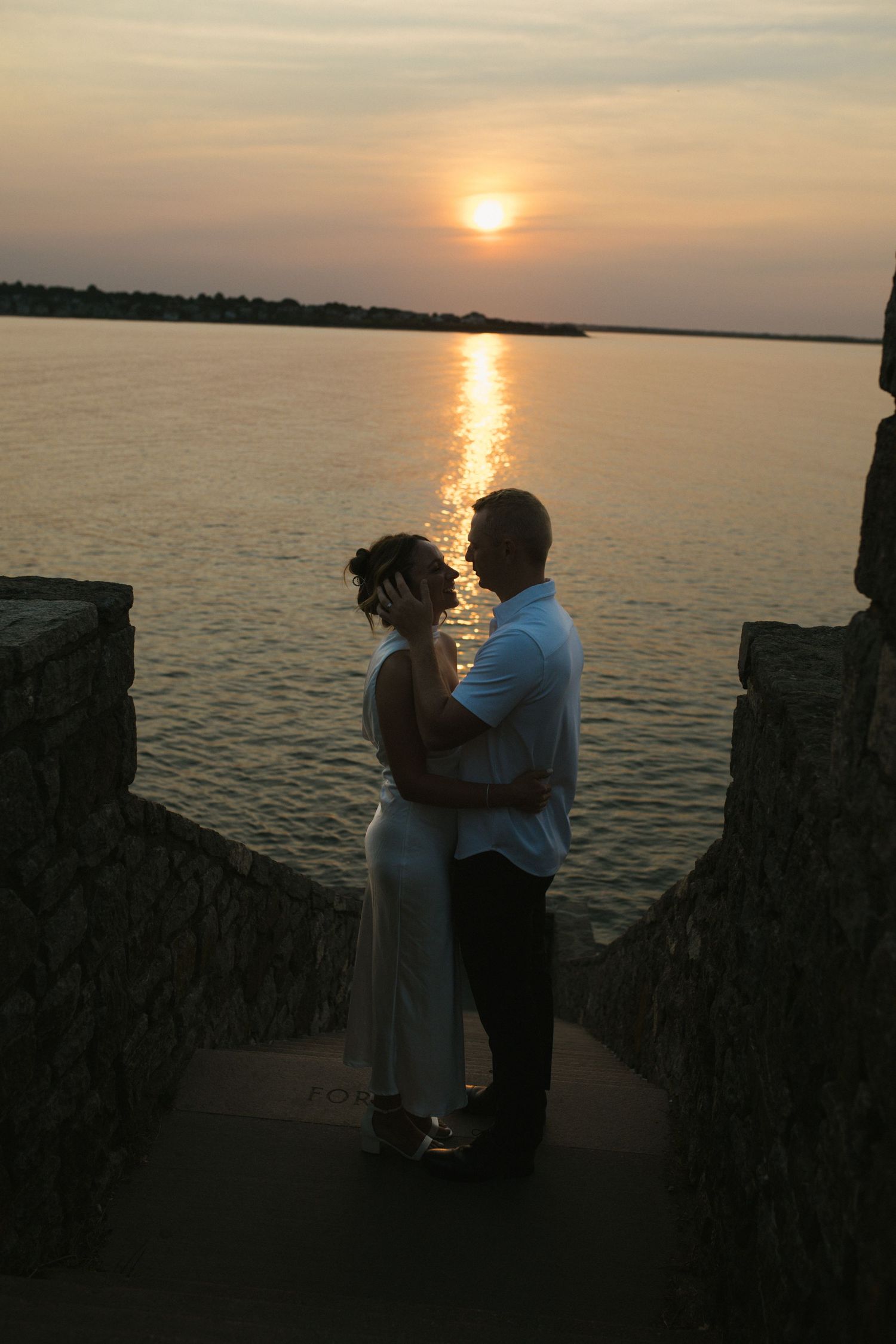 Couple embracing in silhouette between stone pillars at sunset over calm water.
