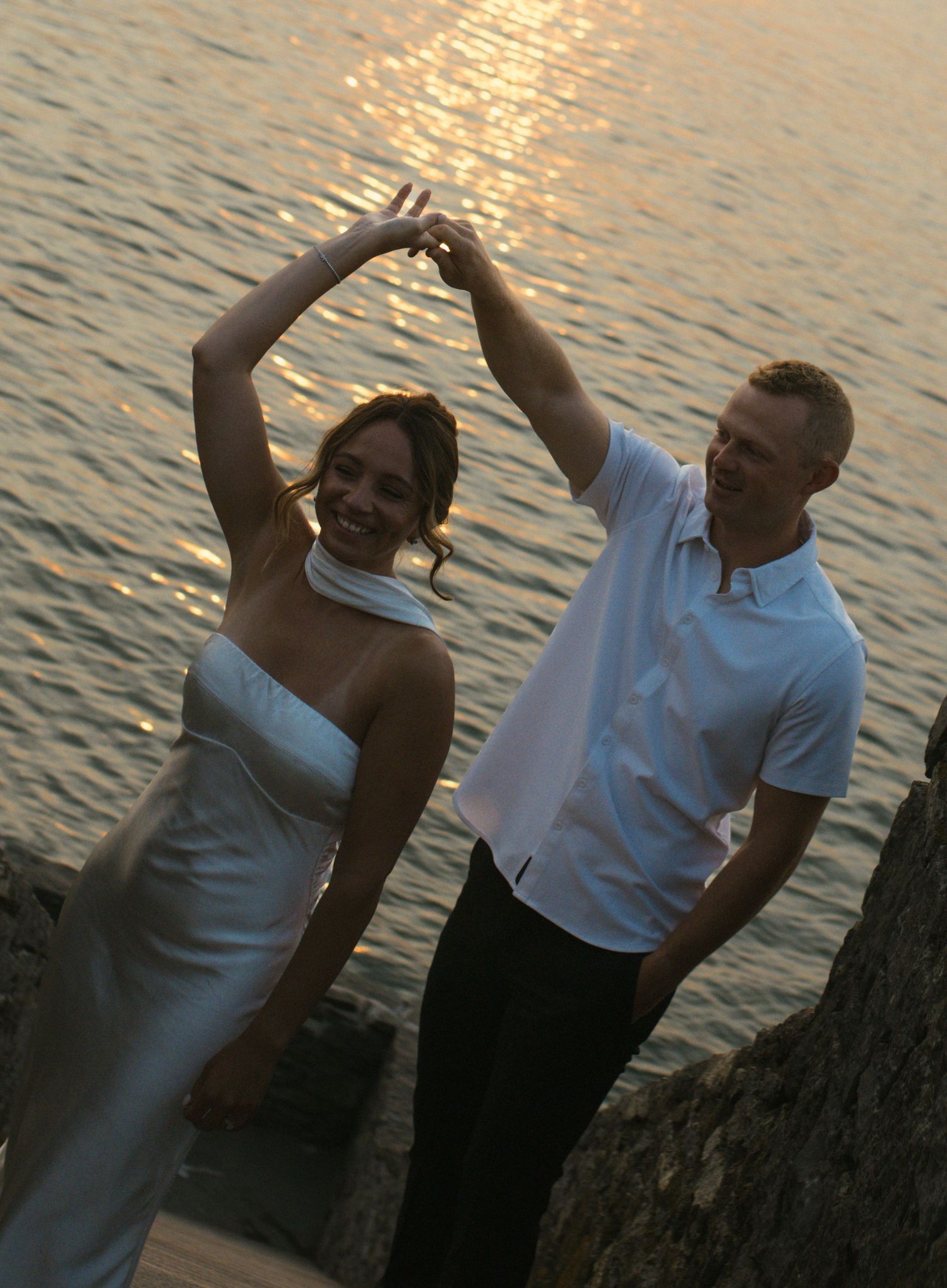 Newlywed couple dancing by the ocean at sunset, bride in white gown, groom in white shirt.