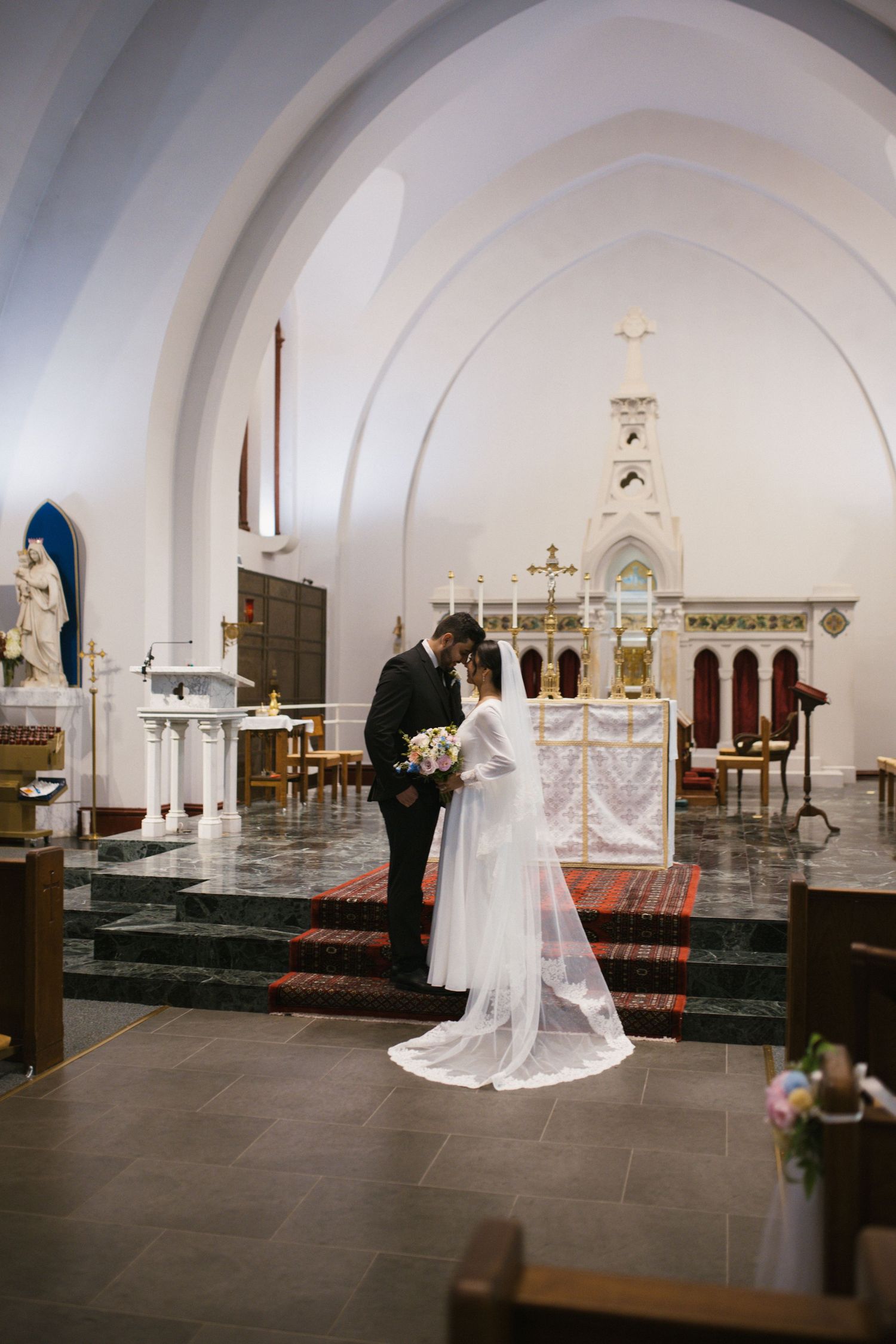 Bride and groom share intimate moment at church altar, with white arched ceiling and ornate decorations in background.