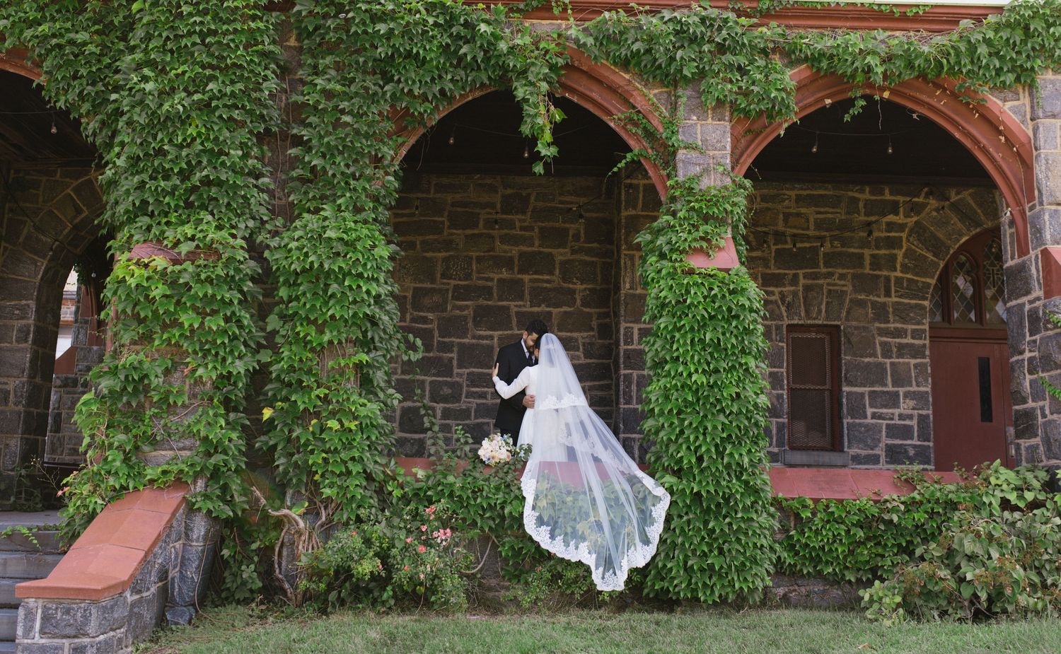 Bride and groom pose by ivy-covered stone arched building, wedding veil flowing in the breeze.
