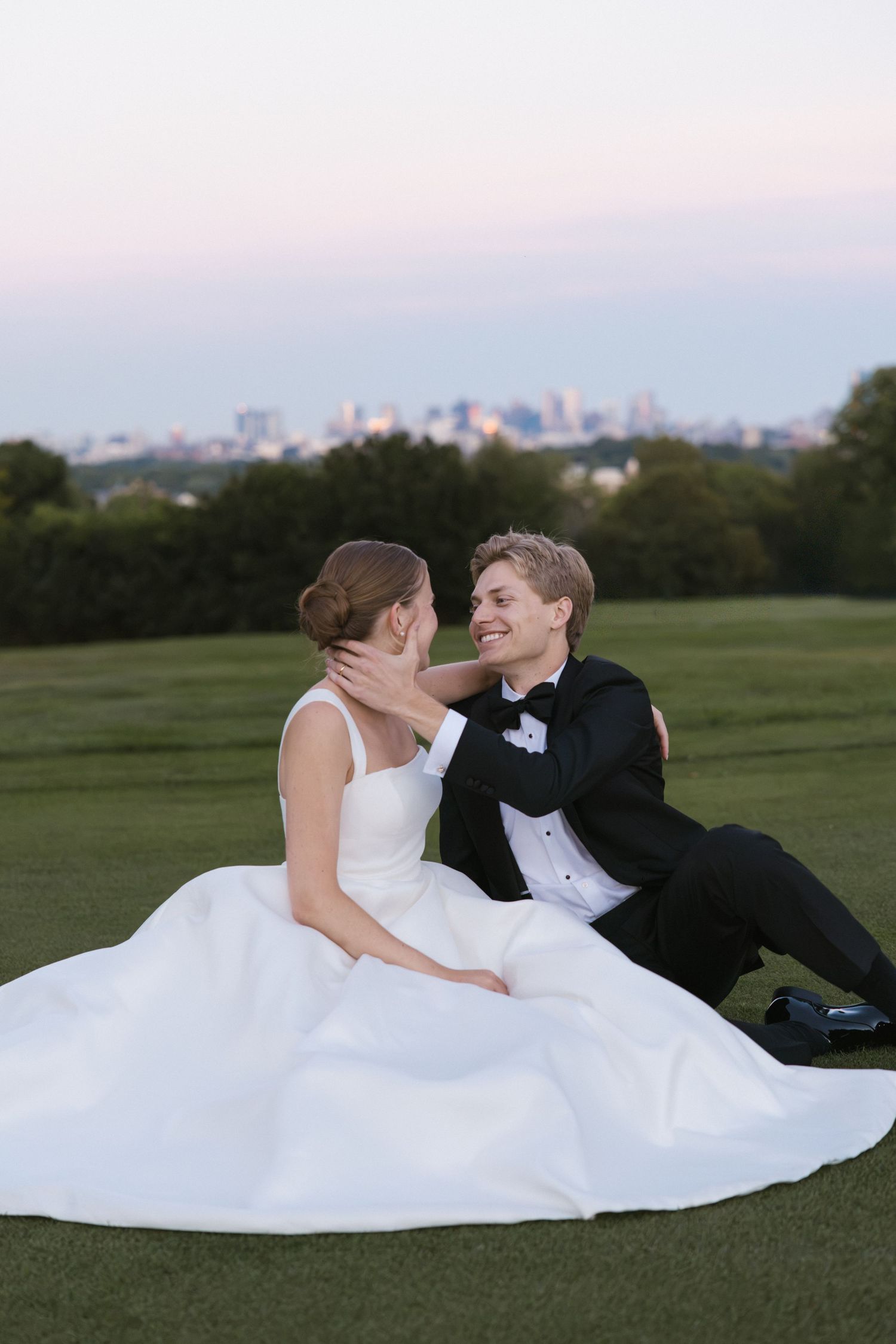 Newlyweds sit together on a green lawn at dusk, groom in tuxedo tenderly holding bride's face with city skyline behind.