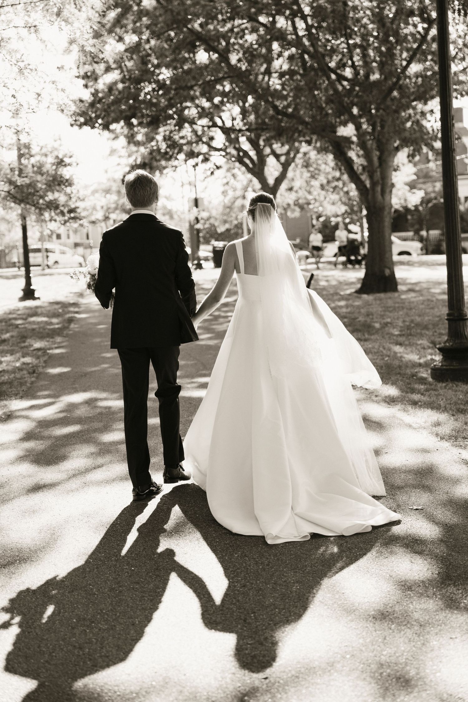 Black and white photo of bride and groom walking hand in hand through a tree-lined park path on their wedding day.