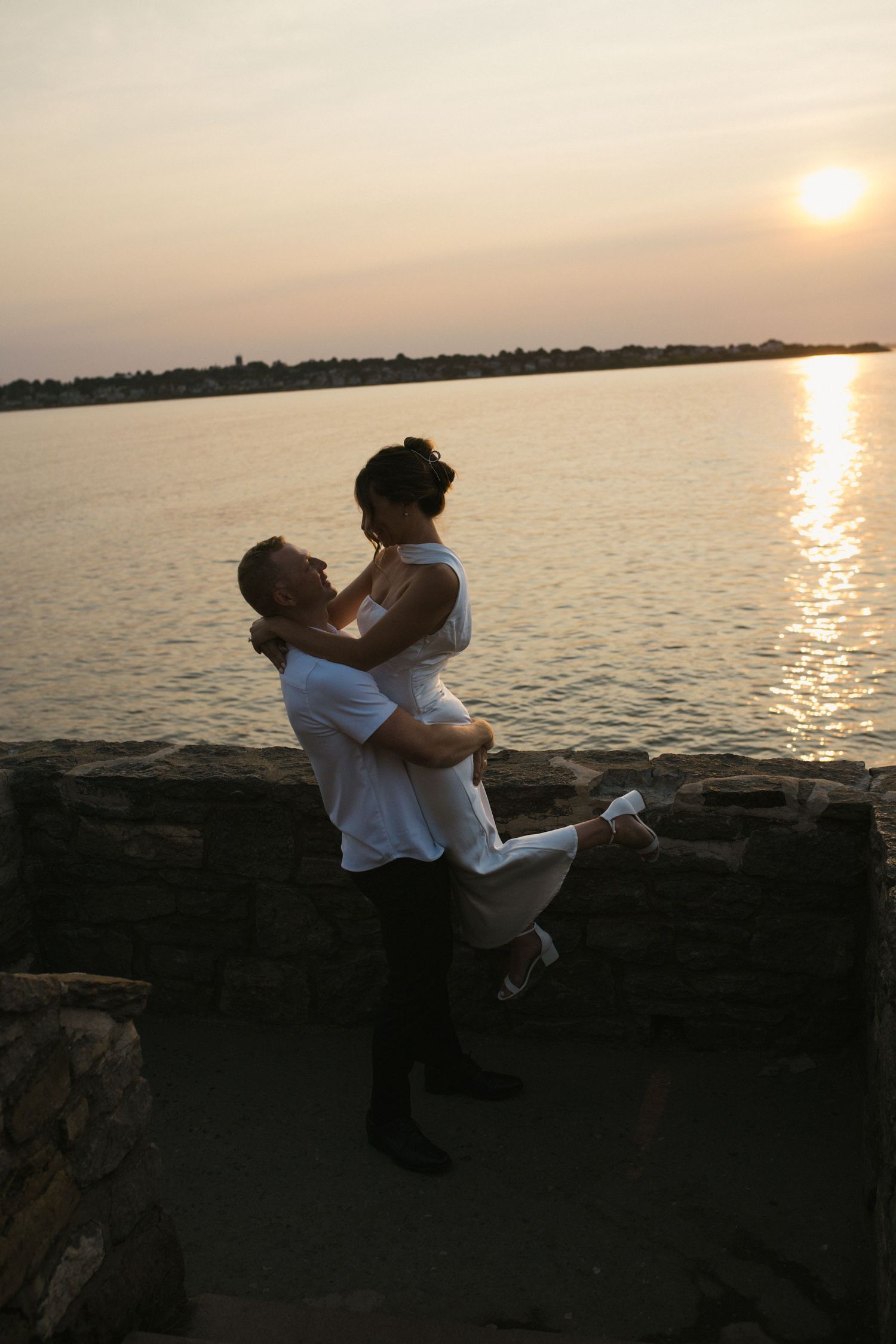 Man lifting woman in white dress at sunset by the water, sharing a joyful romantic moment on rocky shore.