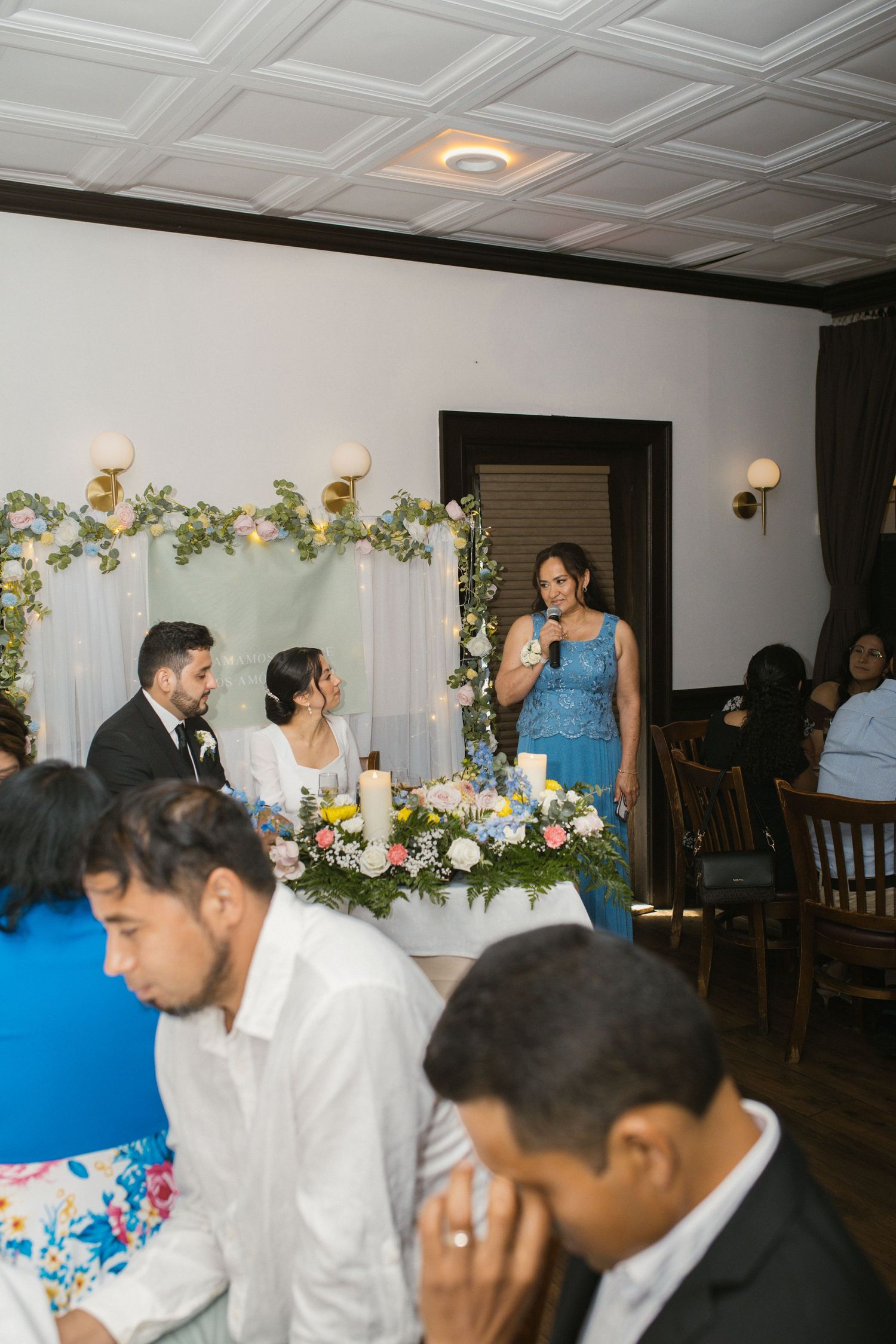 Wedding guest in blue lace dress giving speech at indoor reception while bride and groom listen.