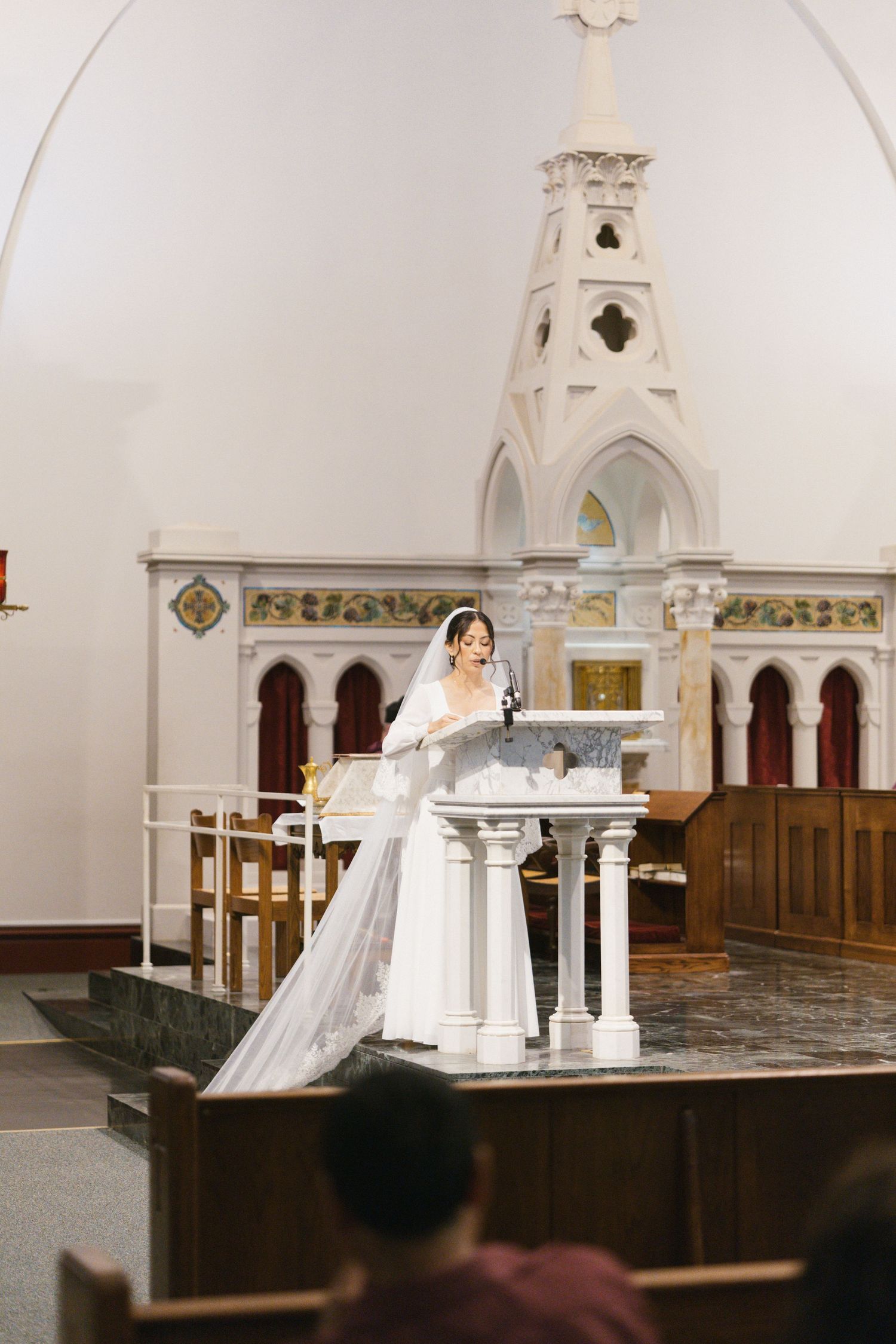 Bride in white gown and veil stands at church altar podium during wedding ceremony.