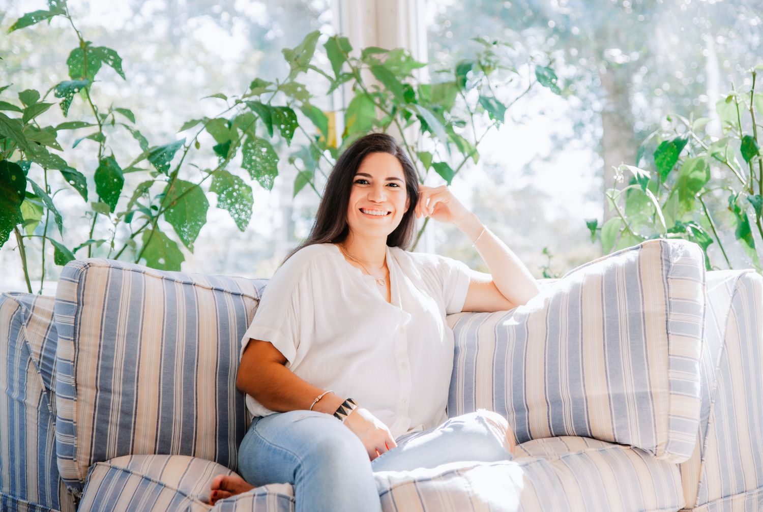 Smiling woman in white top sitting on striped sofa surrounded by green plants.