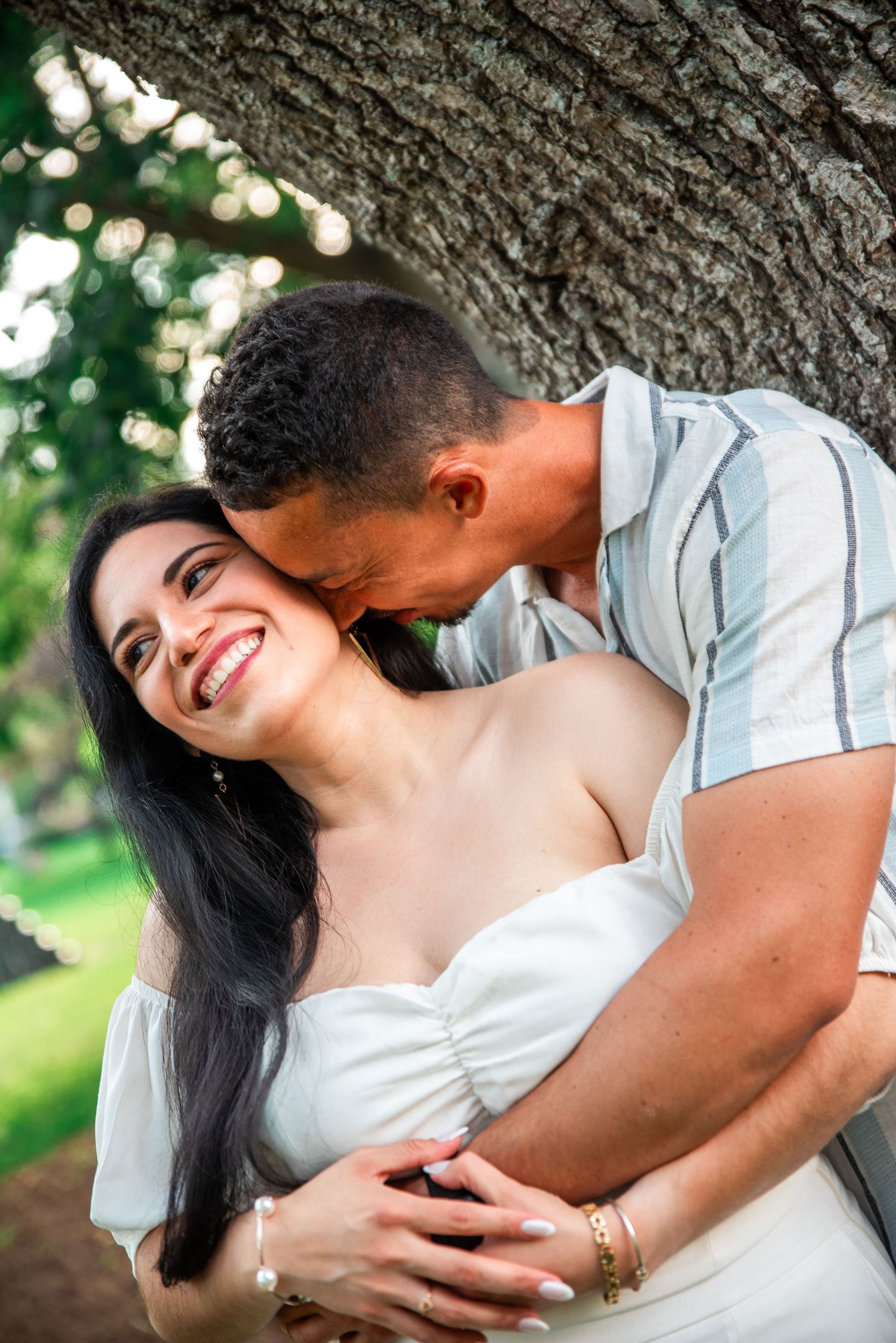 Man kissing smiling woman near tree trunk outdoors in warm sunlight.
