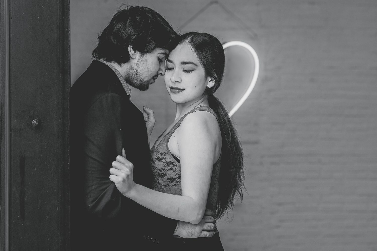 Black and white romantic couple embracing near a heart-shaped neon light in a moody indoor setting.
