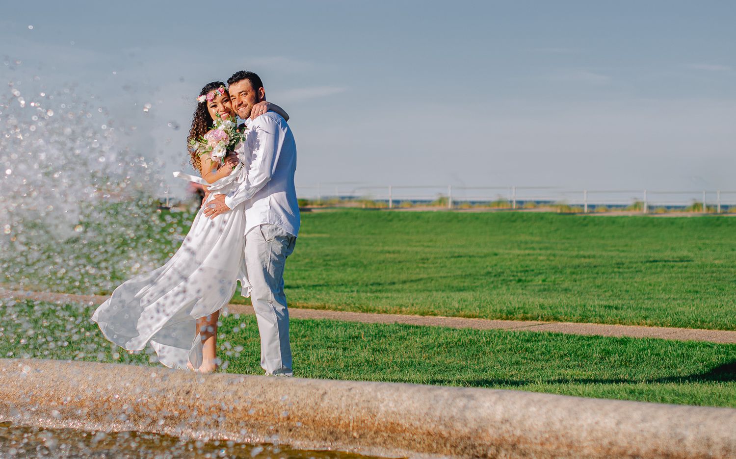 Couple sharing a romantic kiss in wedding attire near a fountain with lush green fields in the background.
