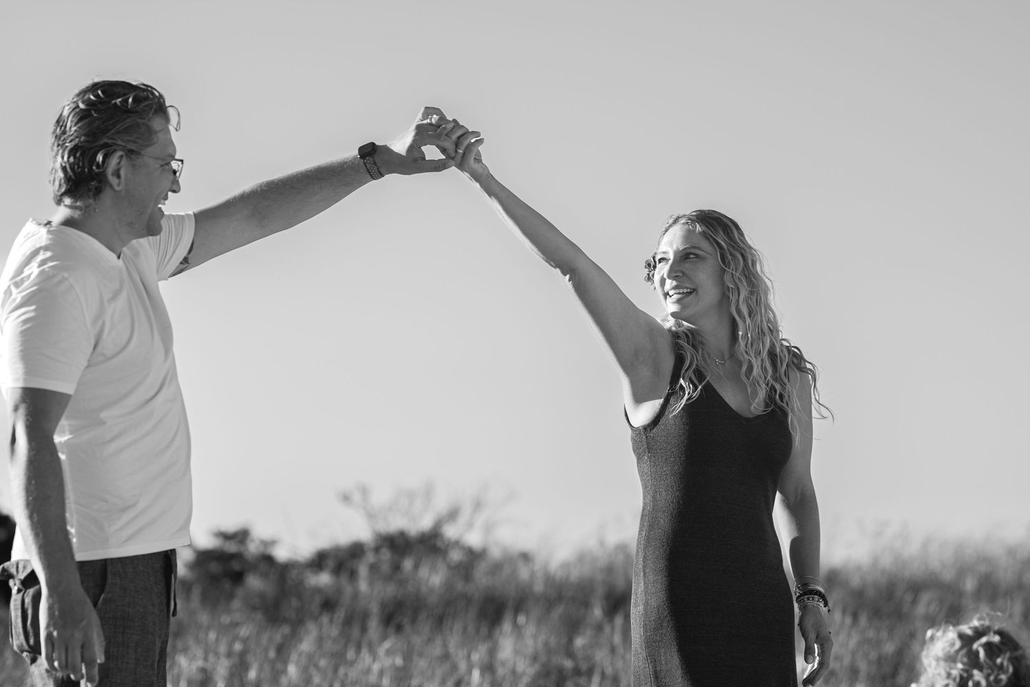 Black and white photo of couple dancing outdoors in field, man spinning woman under raised arms.