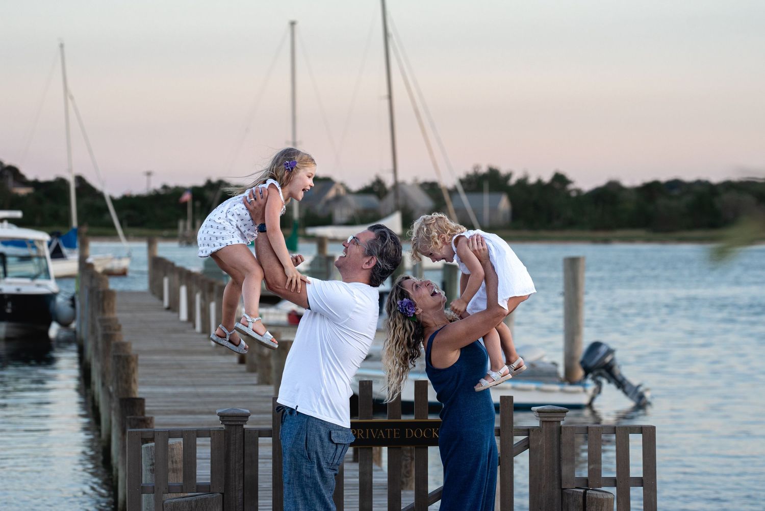 Family of four playing on a wooden dock at sunset with sailboats and calm water in the background. Edgartown lighthouse, Martha's Vineyard