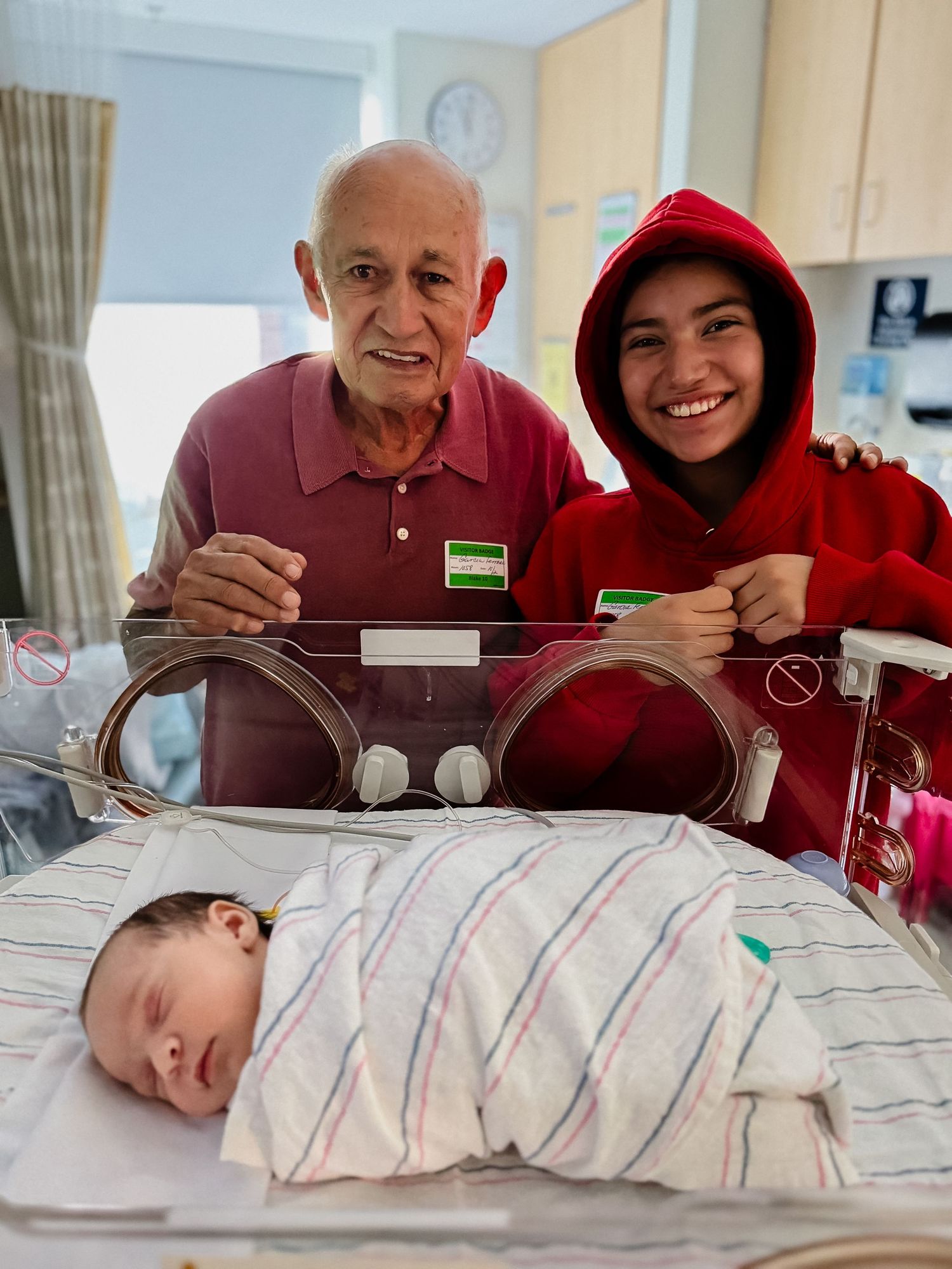 Smiling older man and young person in a red hoodie pose beside a sleeping swaddled newborn in a hospital bassinet.