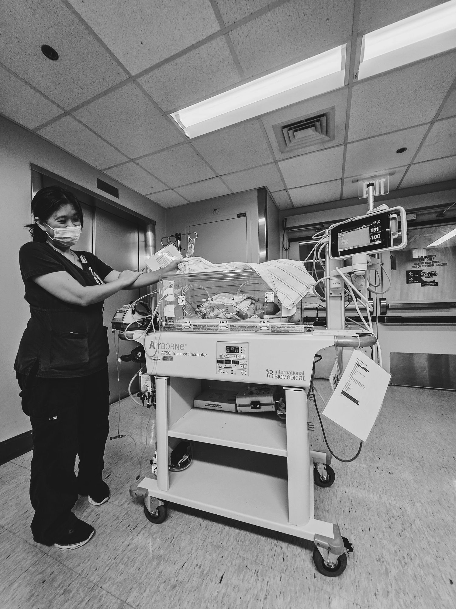 Masked nurse in dark scrubs tends to a newborn in a hospital incubator on a rolling cart in a neonatal care unit.
