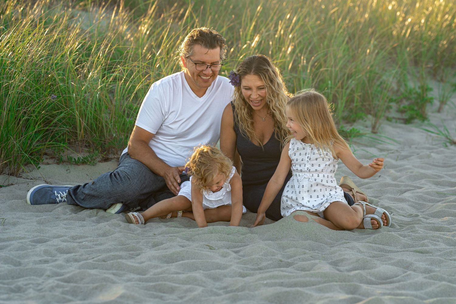 Family of four sitting on sandy beach dunes, parents smiling with two young children, golden hour lighting.