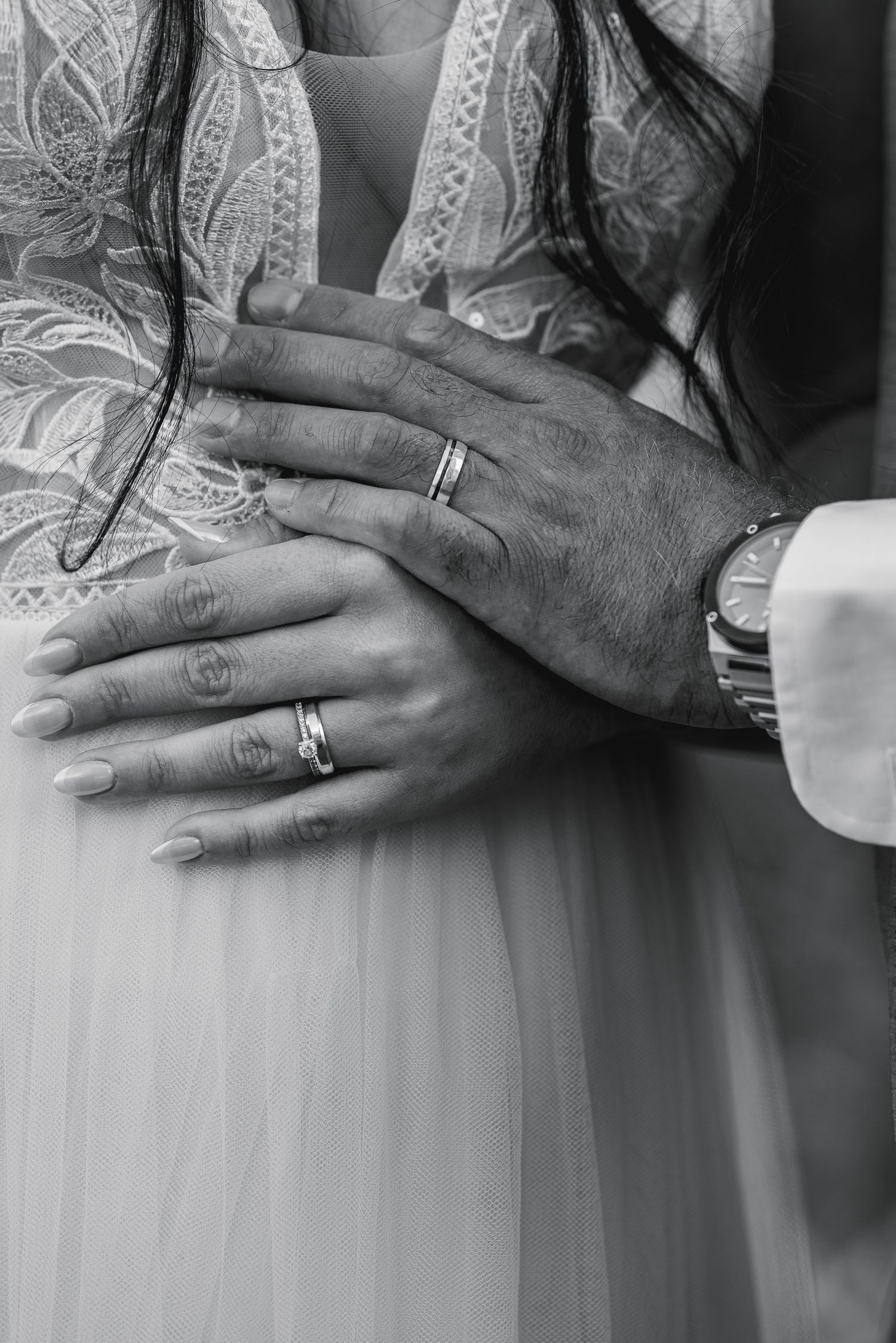 Black and white photo of newlyweds hands with wedding rings, bride in lace dress embraced by groom wearing a watch.