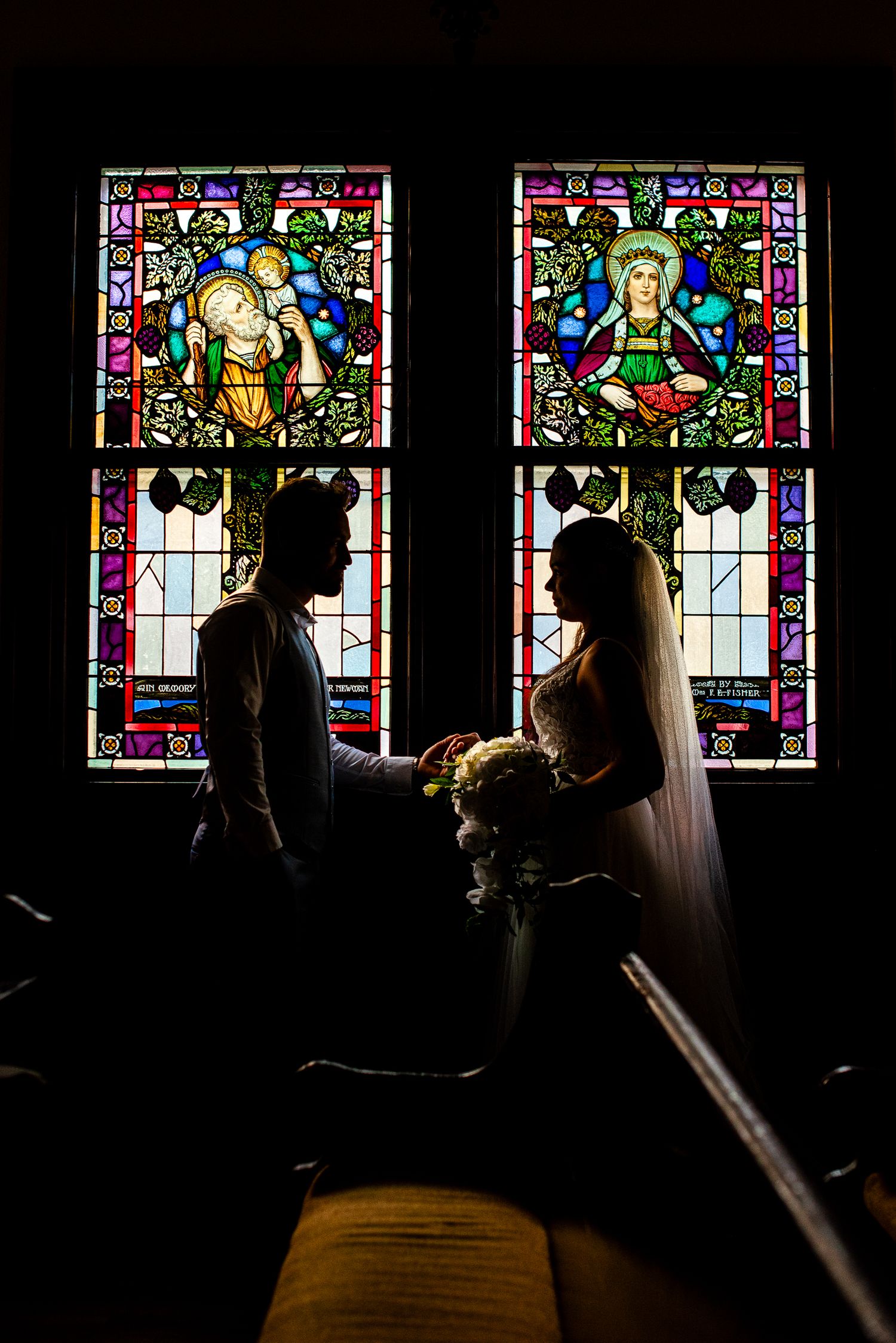 Bride and groom silhouetted against colorful stained glass windows inside a church on their wedding day.
