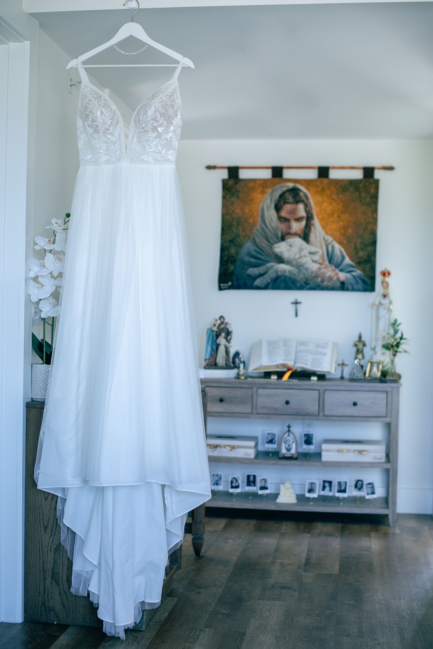 Elegant lace wedding gown hanging near religious artwork of Jesus in bridal preparation room.