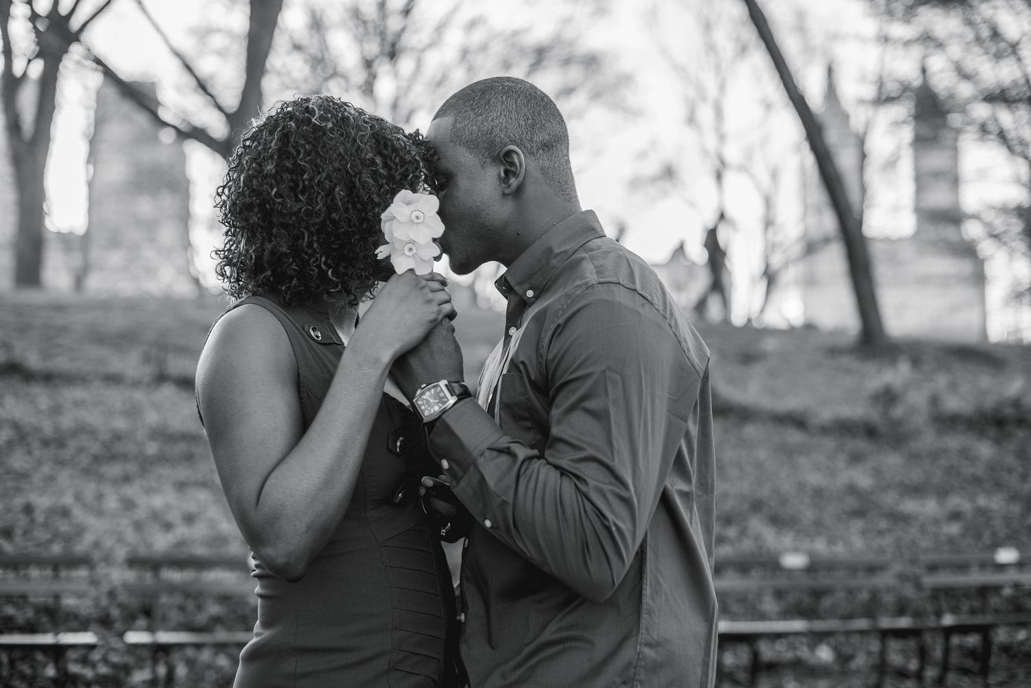 Black and white photo of a couple sharing a kiss behind a white flower bouquet in a park.
