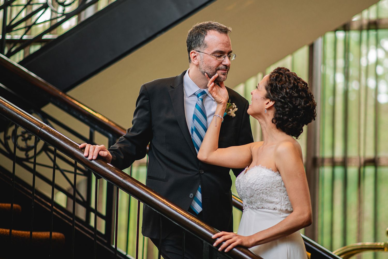Bride and groom share a tender moment on an elegant staircase, with the bride gently touching the groom's face.