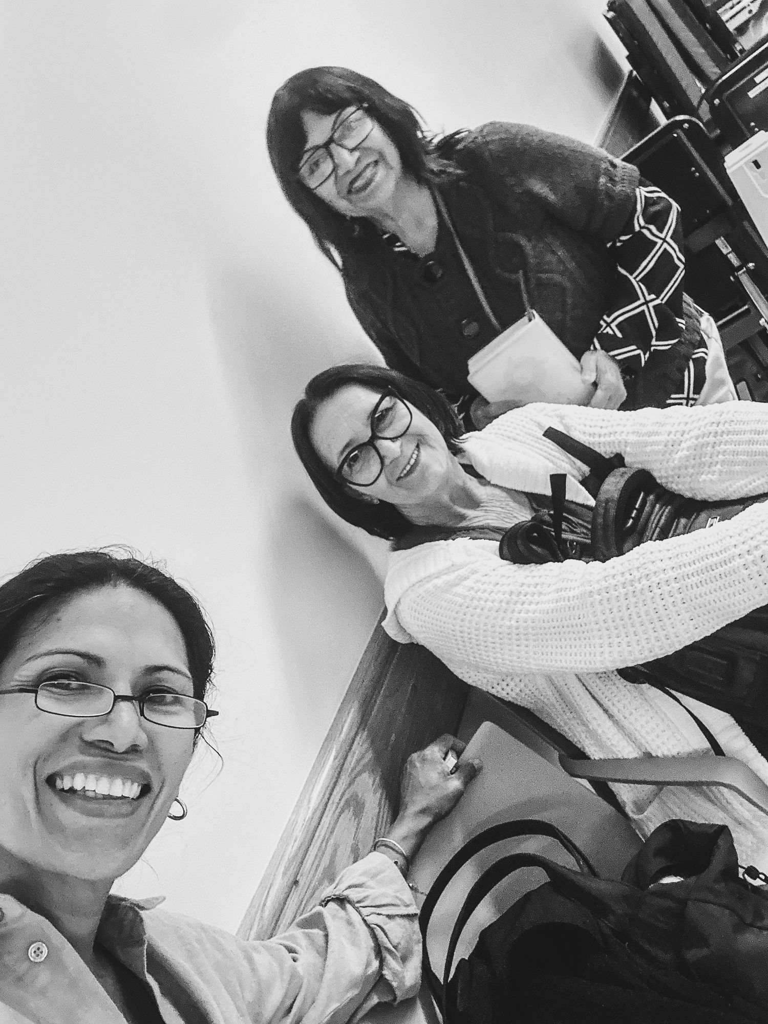 Black and white selfie of three smiling women wearing glasses, one in the foreground and two seated behind her, with bags nearby.