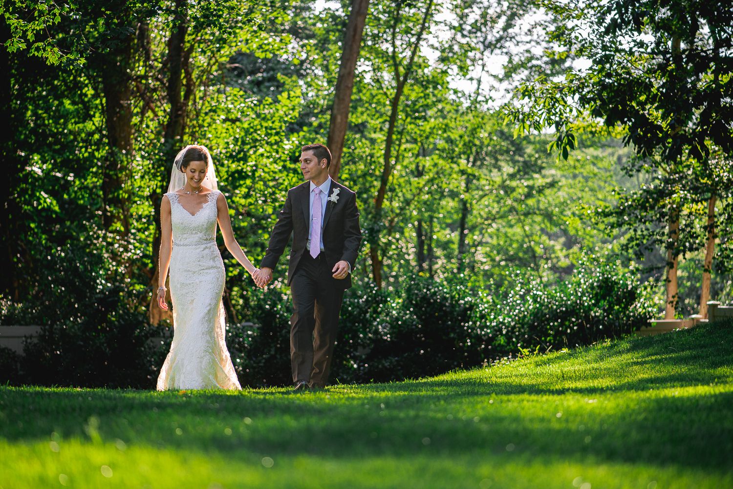 Bride and groom holding hands walking through lush green garden on sunny wedding day.