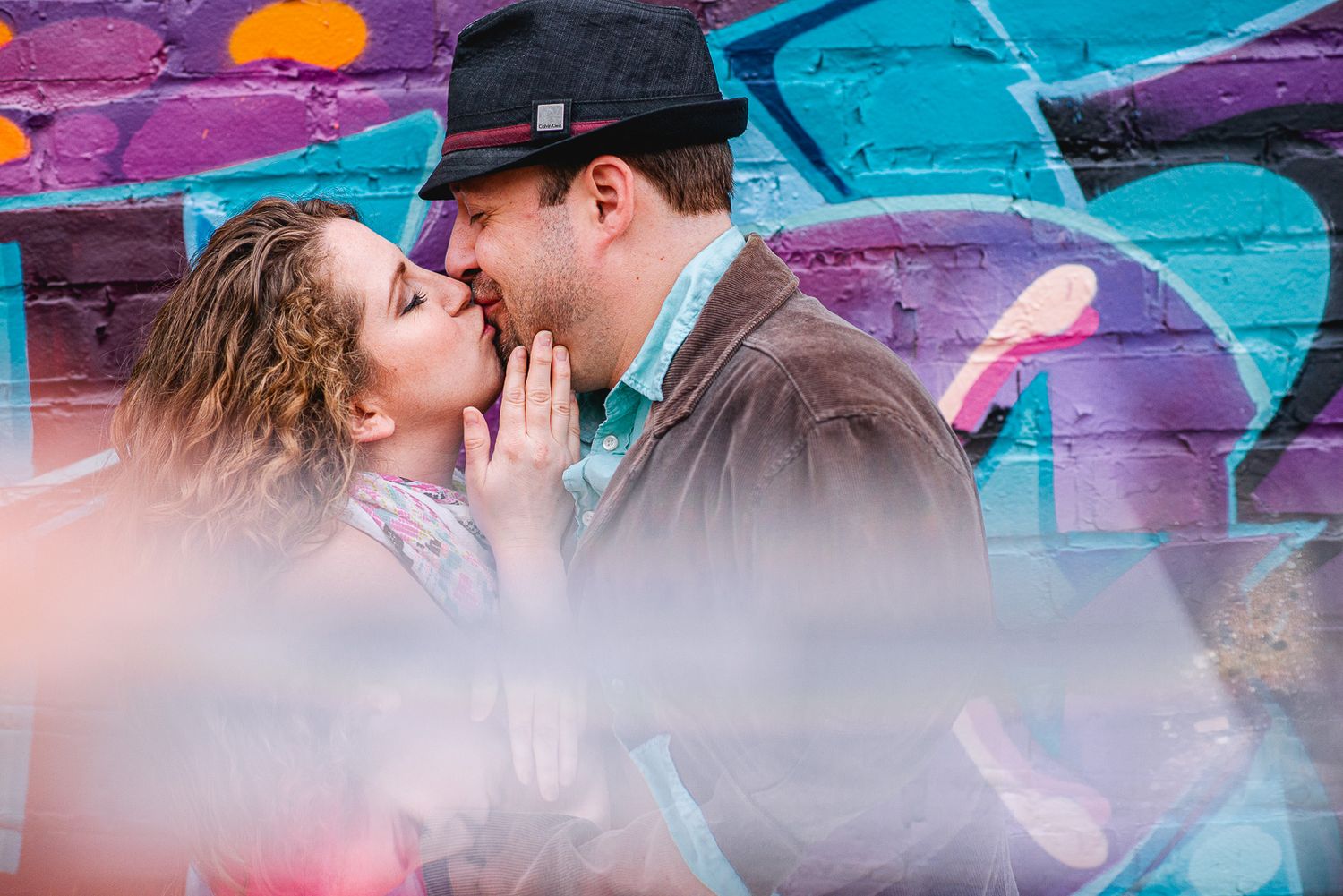 Couple sharing a romantic kiss in front of a vibrant colorful graffiti wall with purple and blue tones.