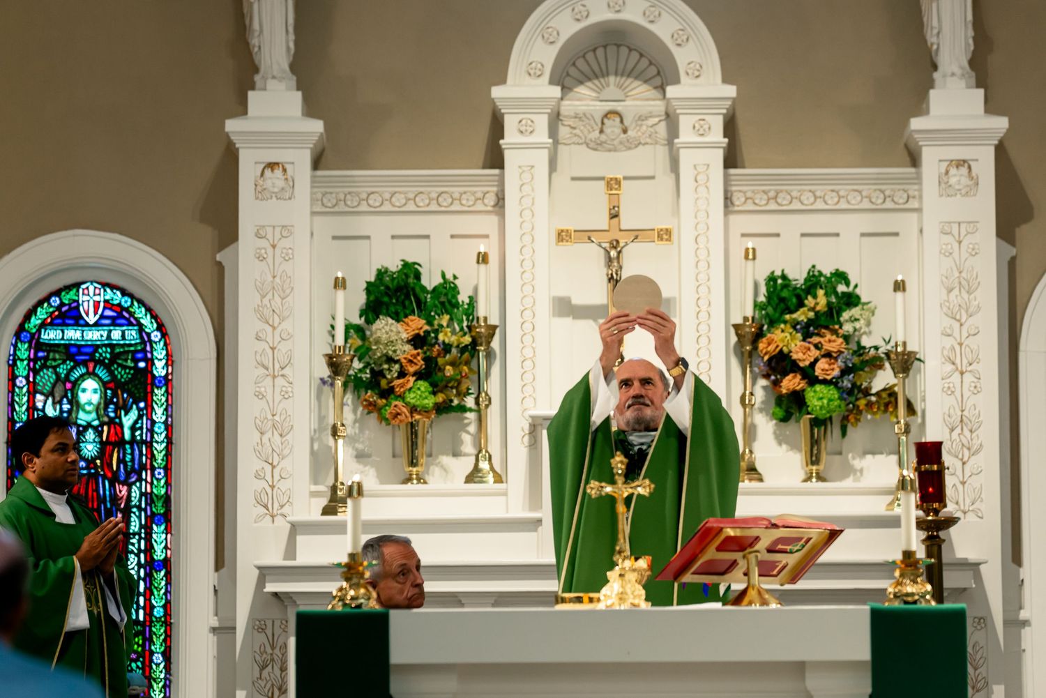 Catholic priest in green vestments elevating the Eucharist during Mass at an ornate white altar with floral arrangements.