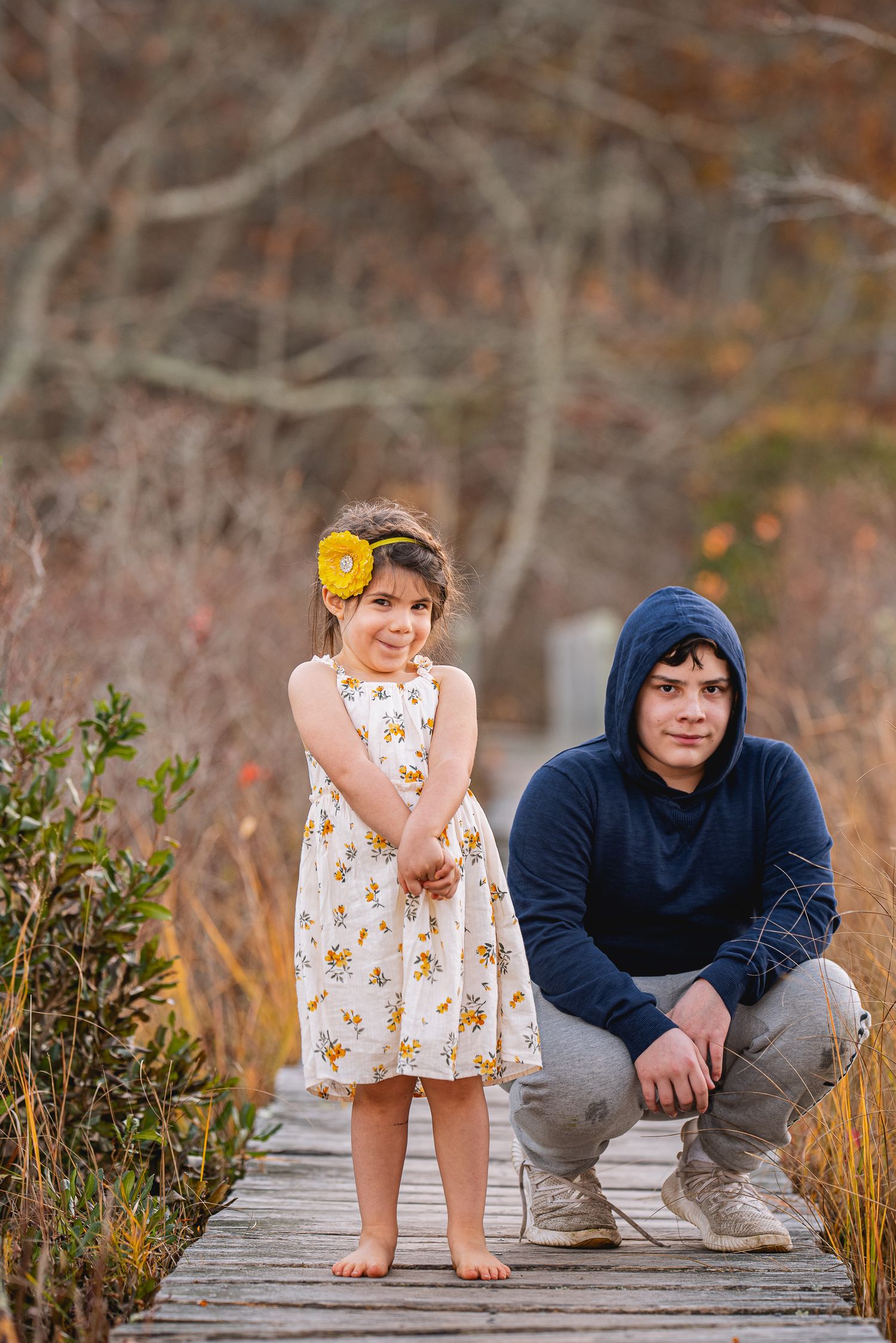 A young girl in a floral dress with yellow flower headband stands beside a teenage boy on a wooden boardwalk in autumn.