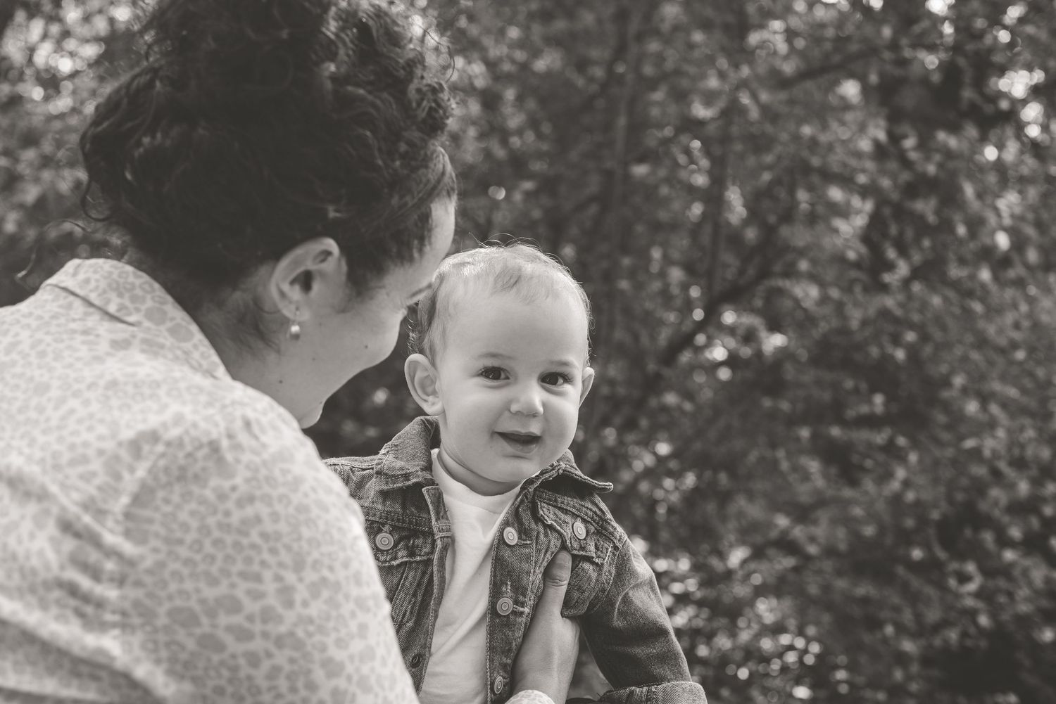 Black and white photo of mother holding smiling baby outdoors with trees in background.