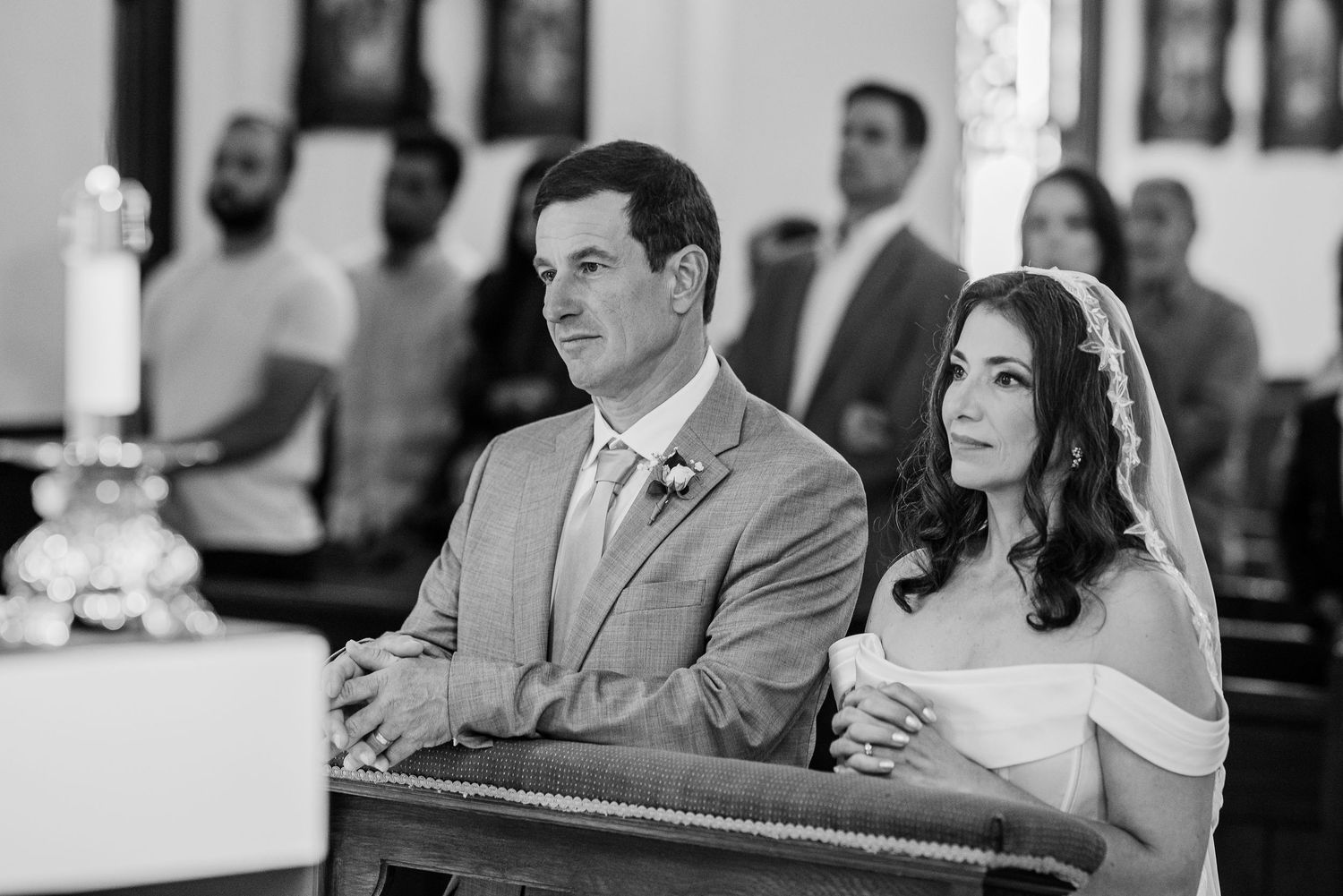 Bride and groom sit together in church pew during wedding ceremony in black and white photo.