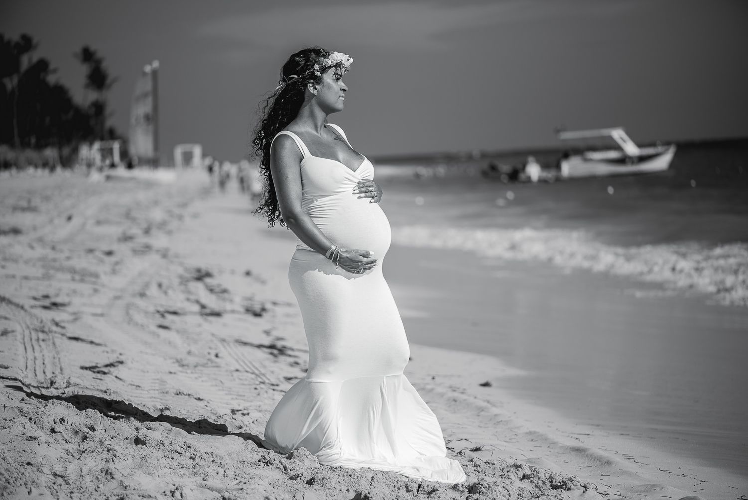 Black and white photo of pregnant woman in white gown on beach wearing flower crown.