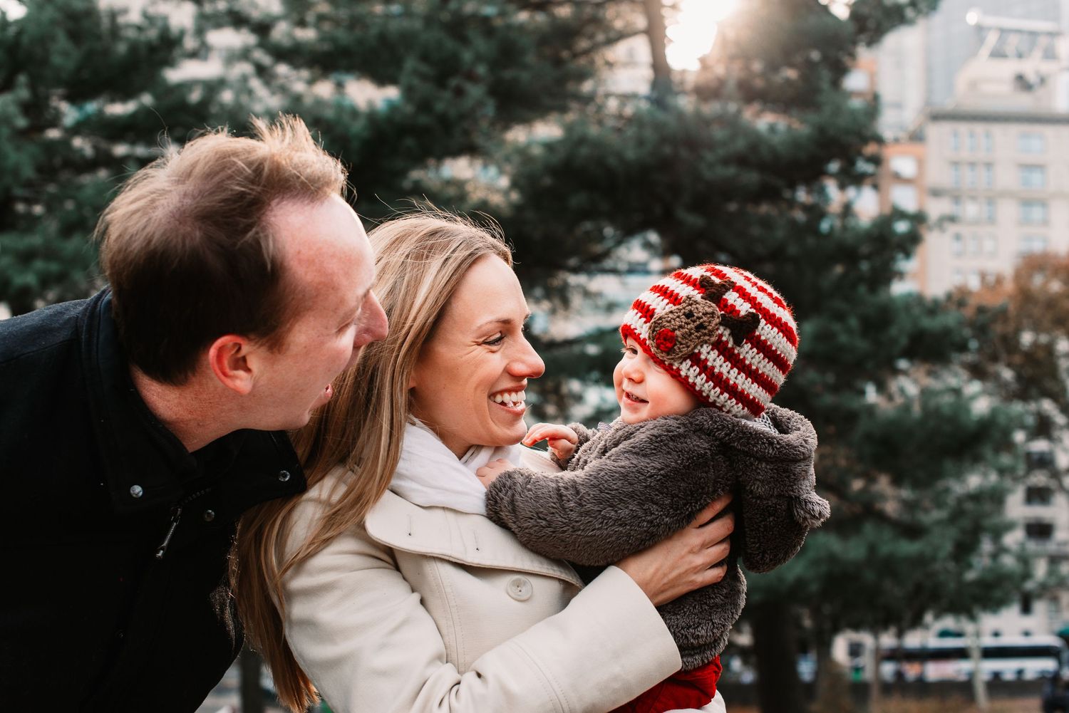 Joyful family with parents kissing and hugging baby wearing red striped hat near Christmas tree.