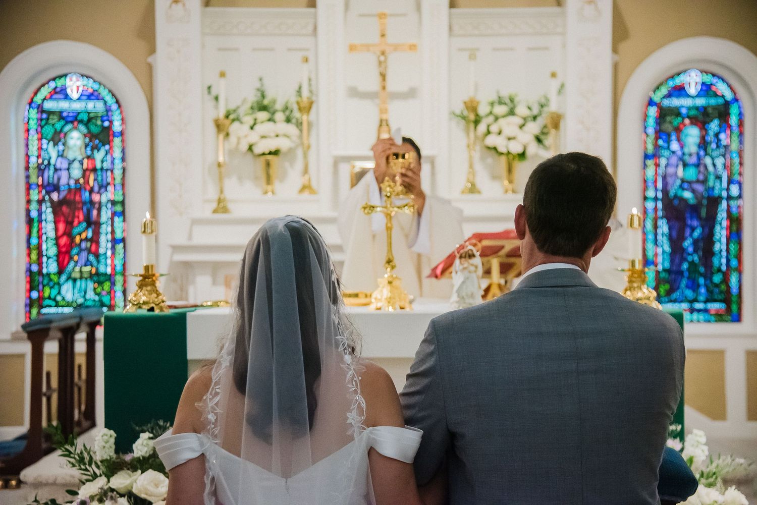 Bride and groom kneeling at church altar during Catholic wedding ceremony with stained glass windows.