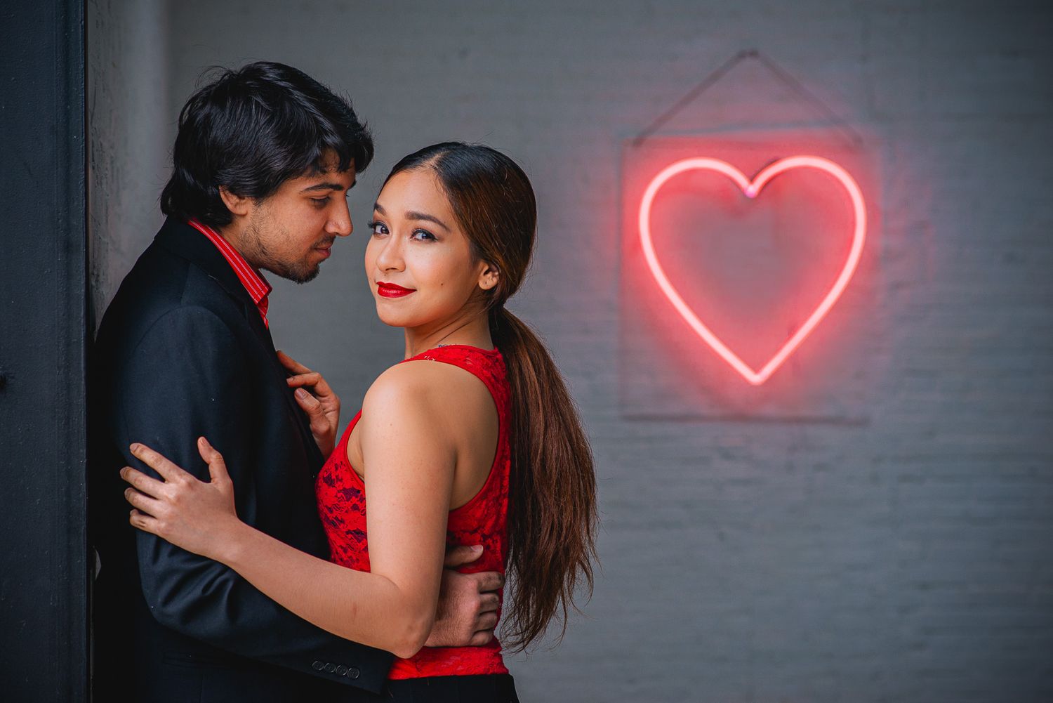Romantic couple embracing near a red neon heart sign, woman in red dress looking back at camera.