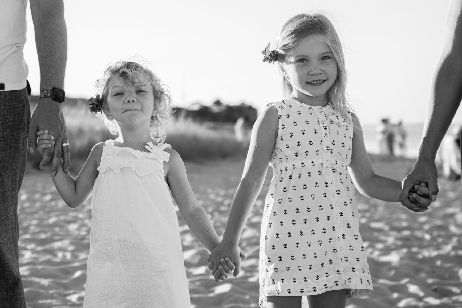 Two young girls in white dresses hold hands on a beach in a black and white photo.
