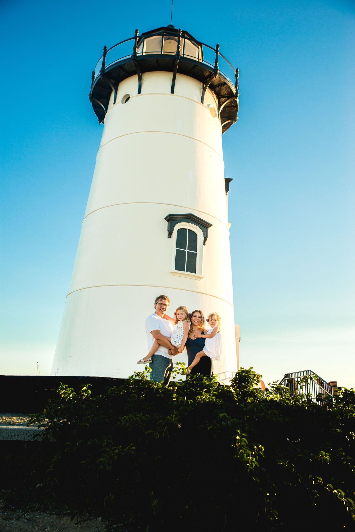Family of four posing in front of a white lighthouse under a clear blue sky on a sunny day.